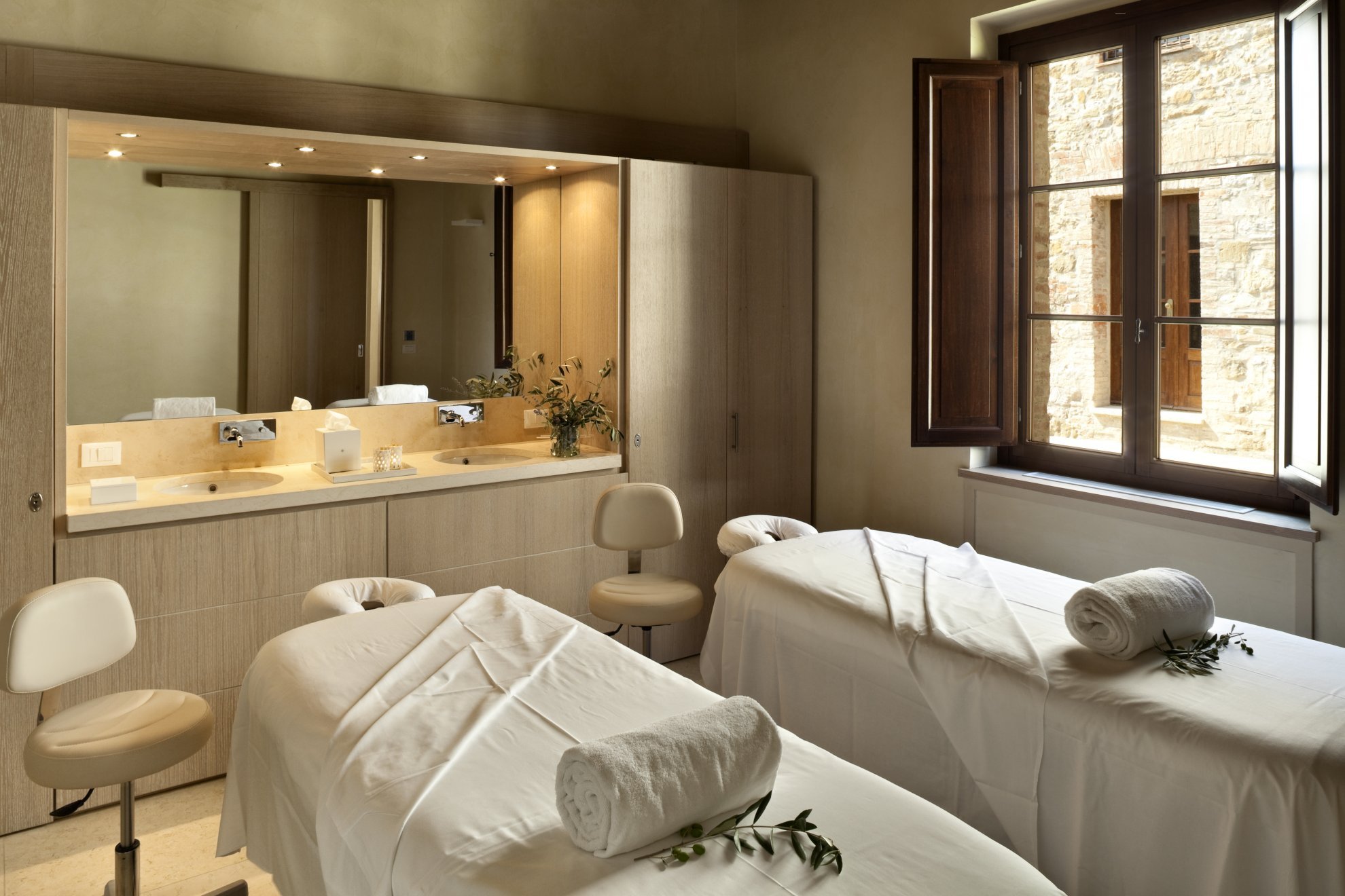 Cozy spa treatment room with two massage beds adorned with white towels and plants. A large window lets in natural light, illuminating the soft, neutral tones.