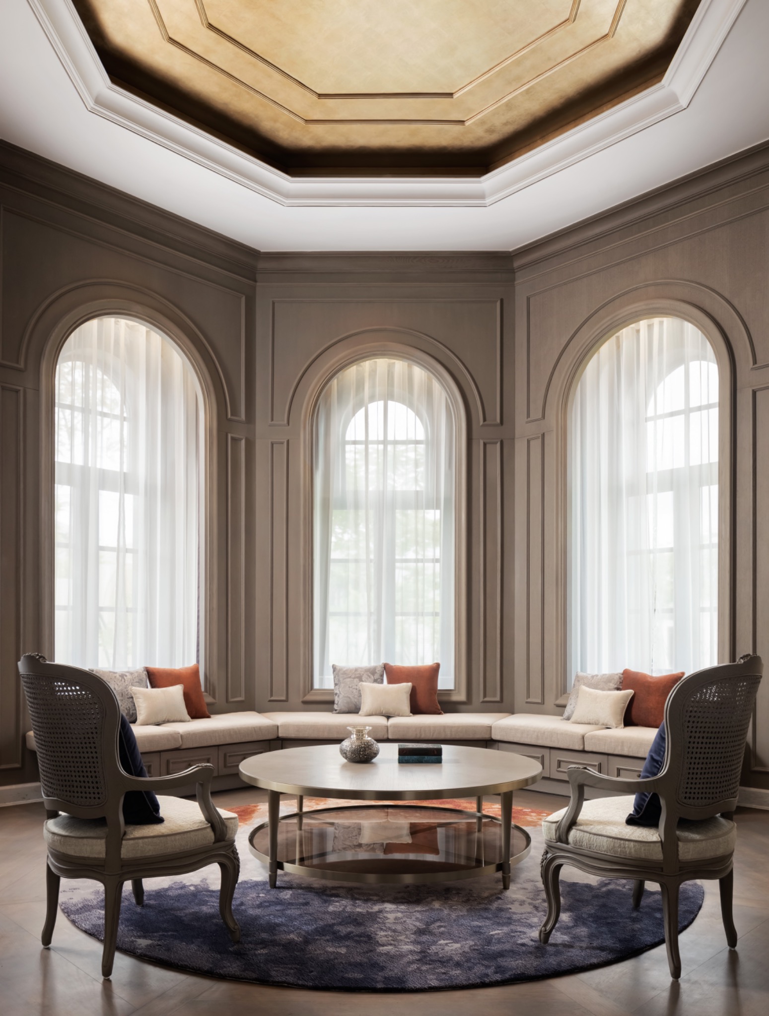 Elegant living room with high, coffered ceiling and large arched windows. Features round coffee table, cozy beige sofas with colorful pillows, creating a serene ambiance.