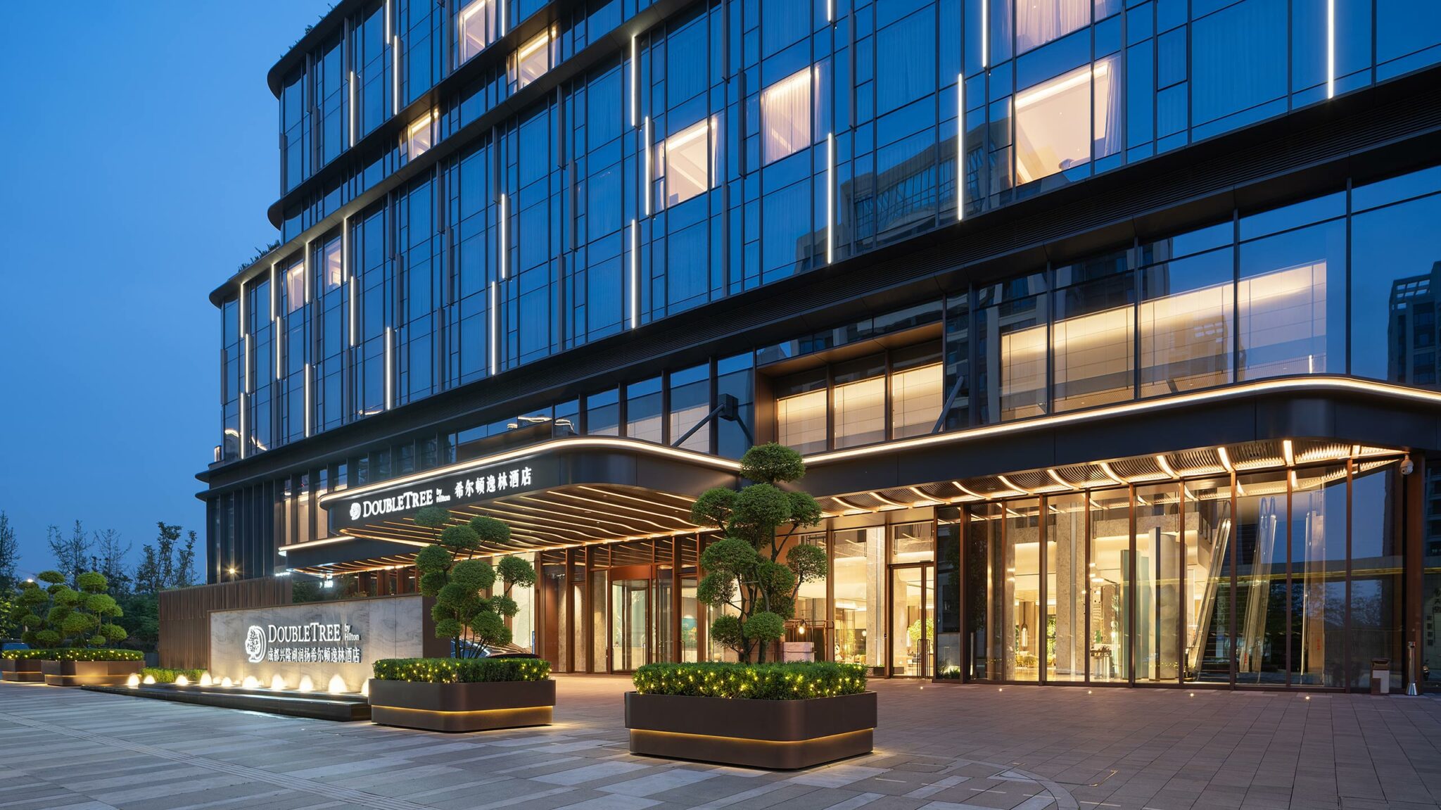 Modern hotel entrance at dusk, featuring large glass facade and warm lighting. Trimmed greenery adds elegance. An inviting and upscale ambiance.