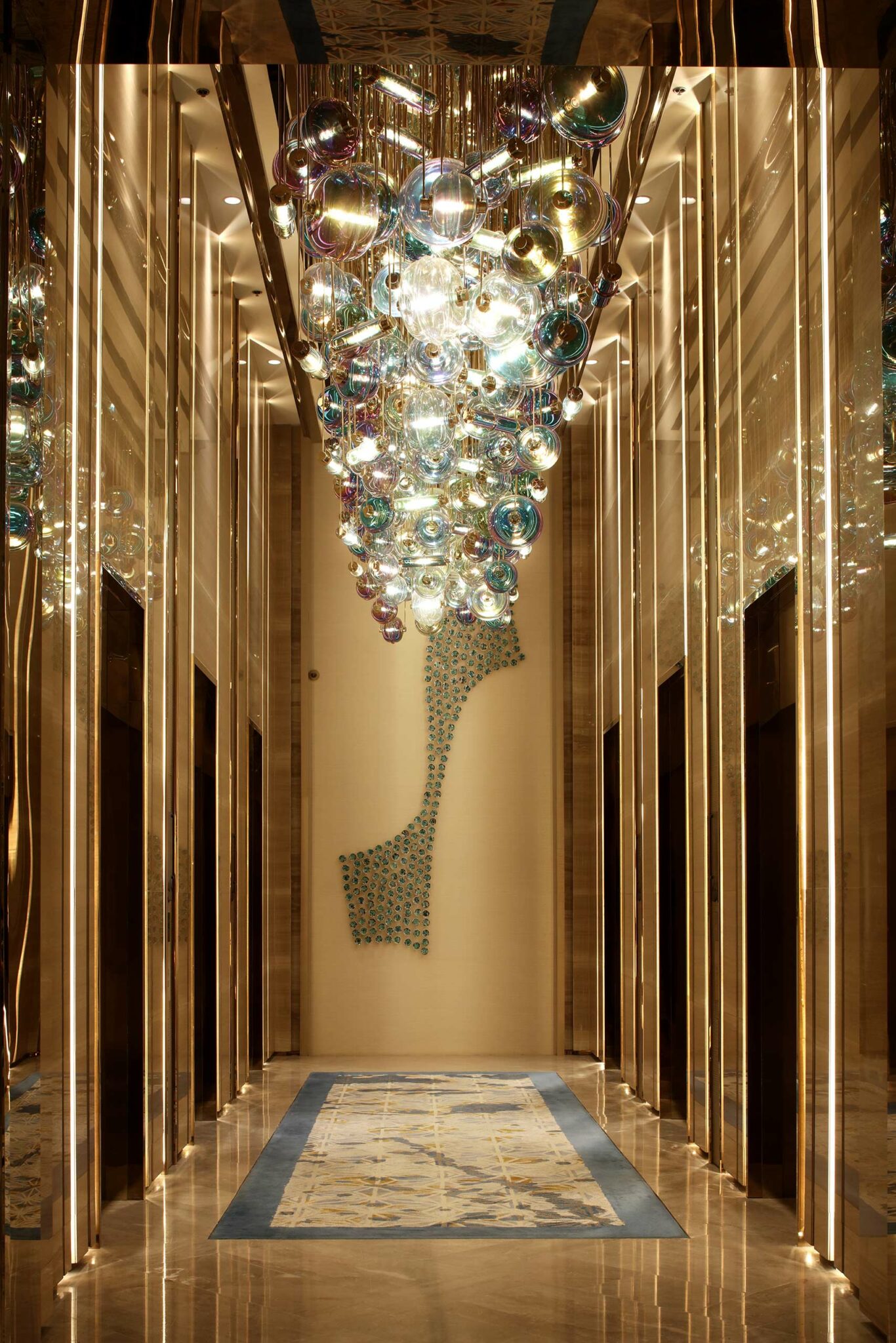 Luxurious hotel hallway with a vibrant, multicolored glass chandelier. Reflective walls enhance the glow, leading to a patterned wall art. Opulent and modern.