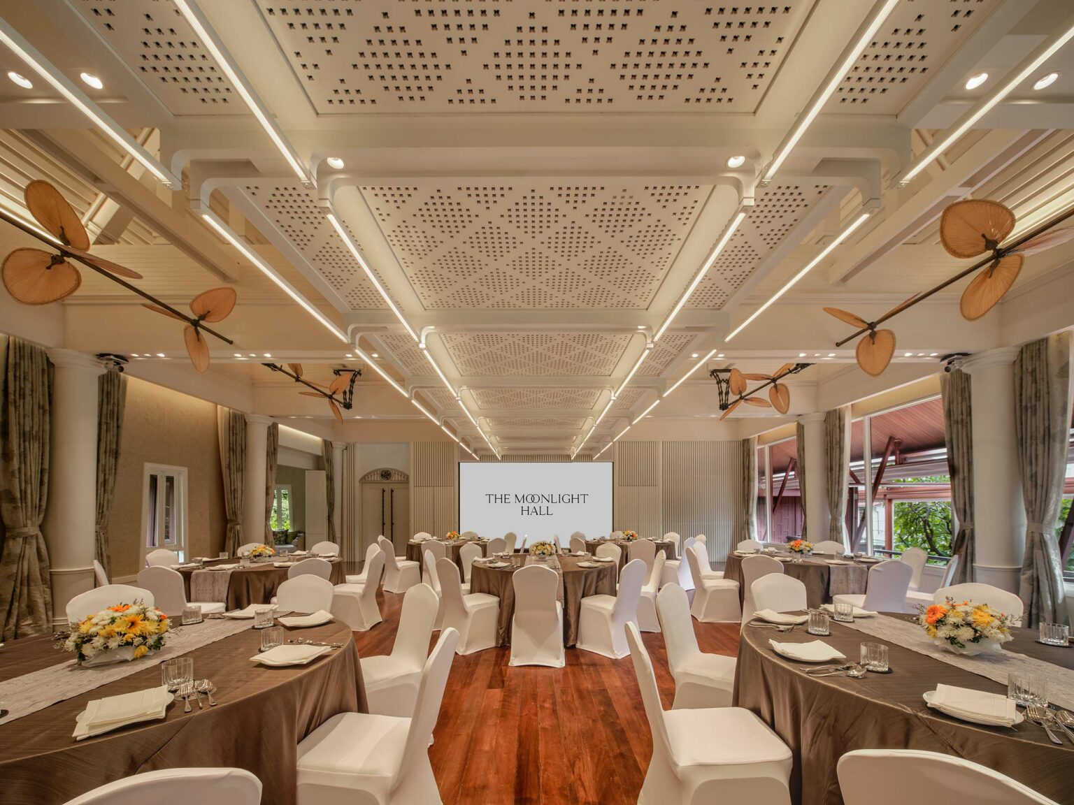 Elegant hotel banquet hall with round tables covered in brown cloth and white chairs. Floral centerpieces add warmth. Ceiling fans and modern lighting create a sophisticated ambiance.