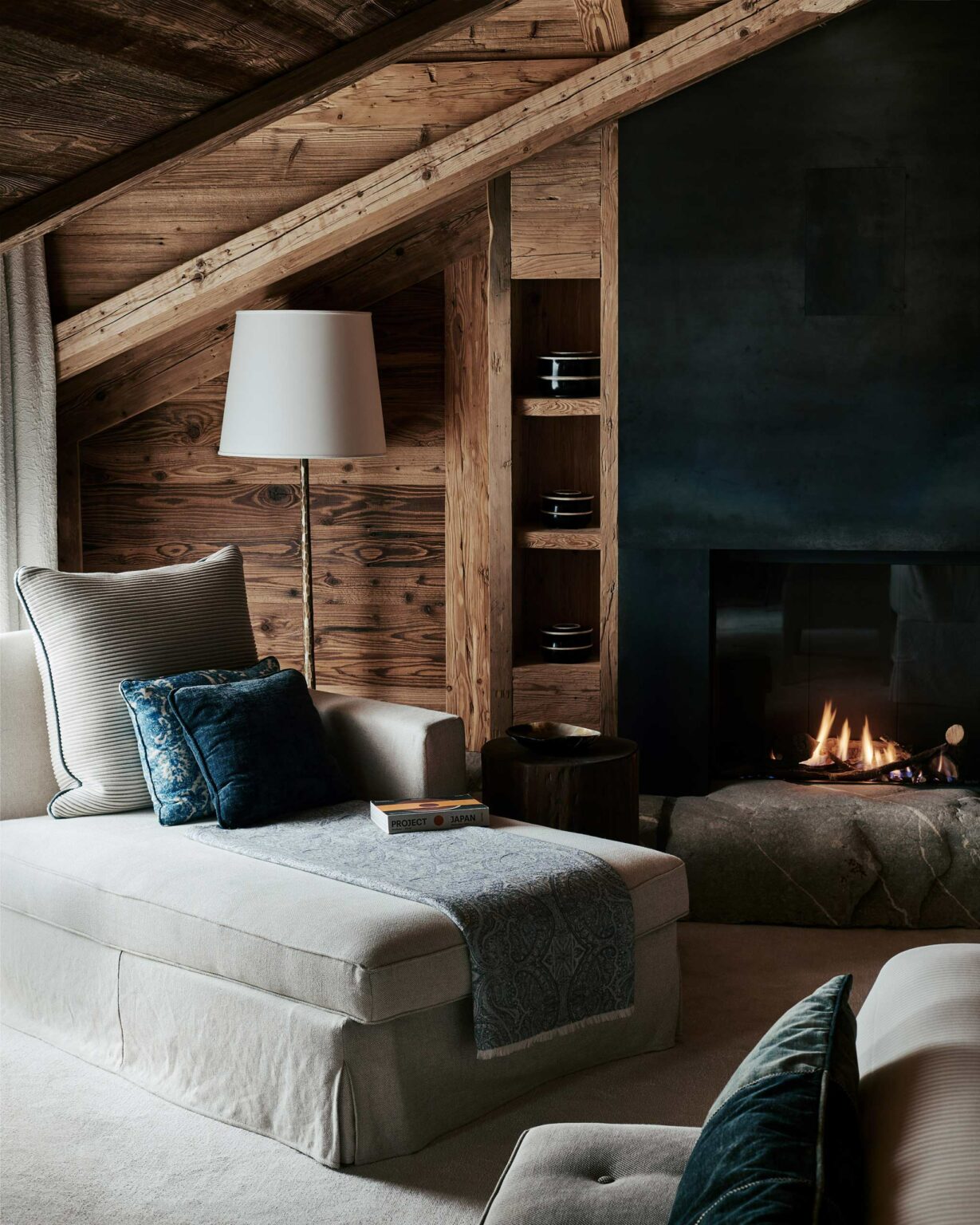 Cozy rustic nook of a guest room featuring a sloped wooden ceiling, cream chaise longue, blue cushions, a stone fireplace with a small fire, and books on the side table.