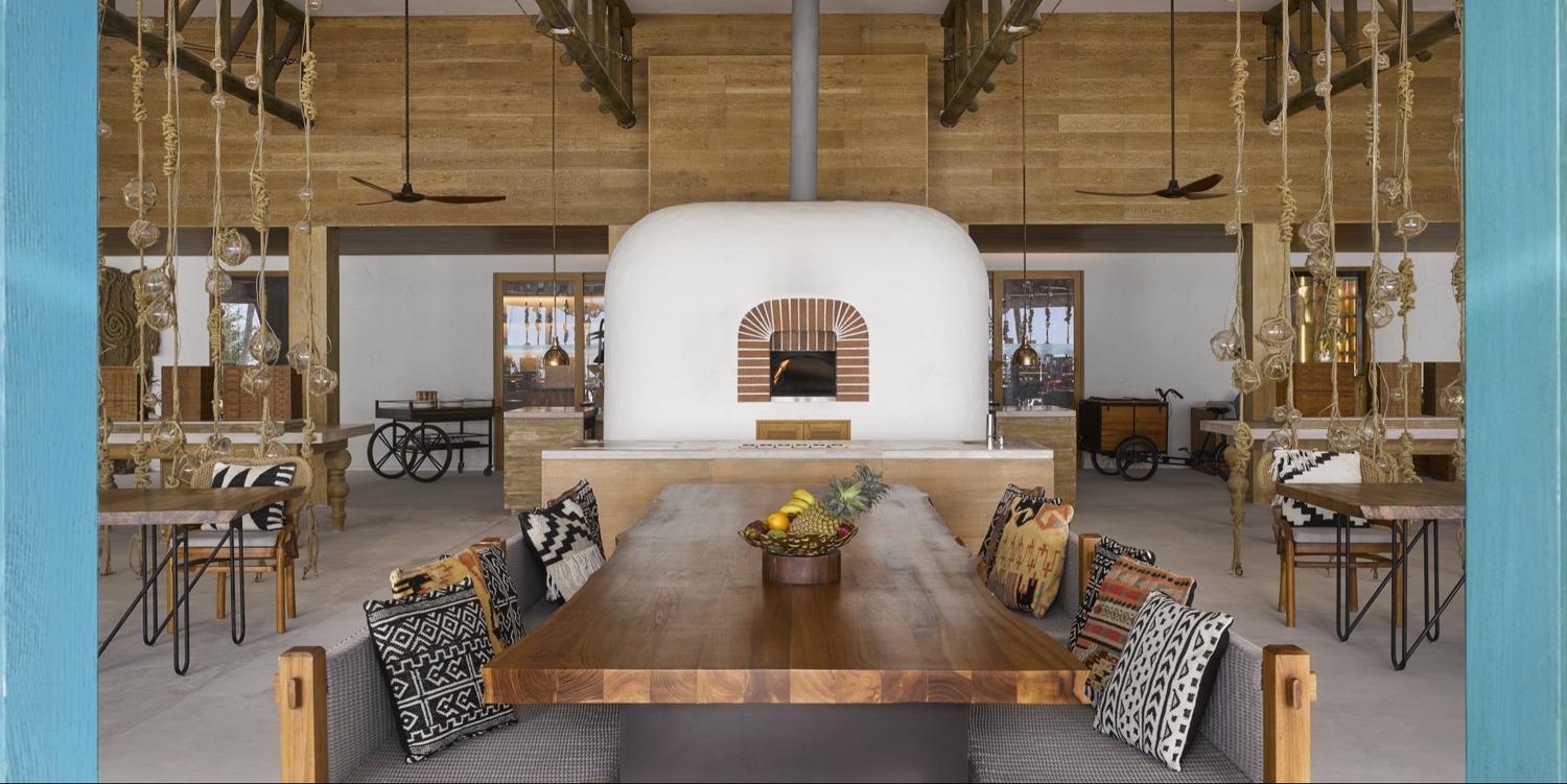 Cozy beachside resort restaurant interior with warm lighting, woven ceiling, and rustic decor. Open walls reveal an ocean view at sunset, creating a serene atmosphere.