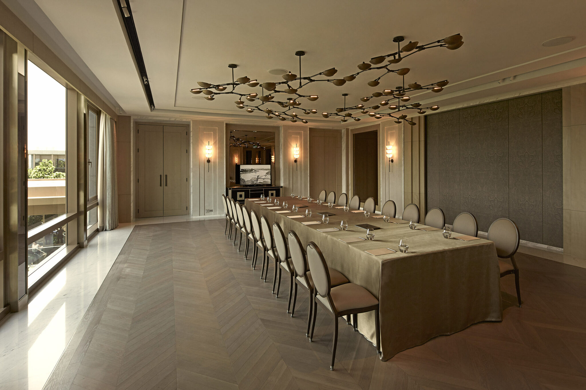 Spacious, elegant hotel conference room with a long table, beige chairs, and artistic ceiling lights. Sunlight streams through large windows, creating a warm ambiance.