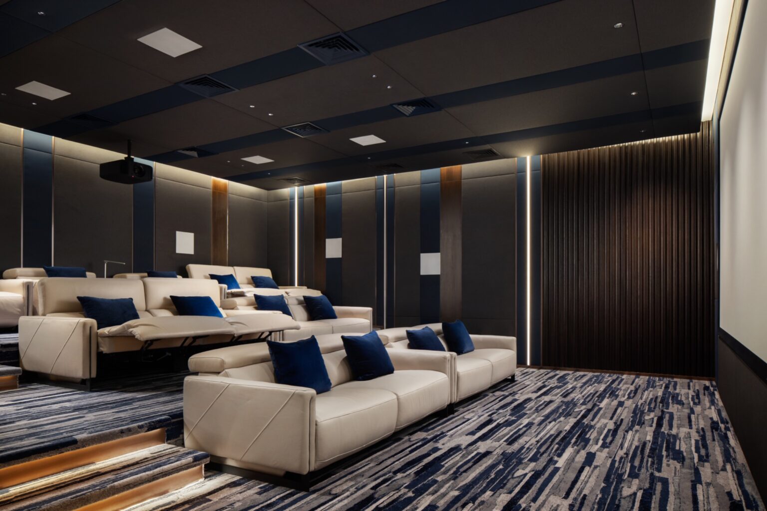 Luxurious home theater with beige recliner sofas, blue cushions, and modern dark walls. A large screen and striped carpet complete the serene setting.