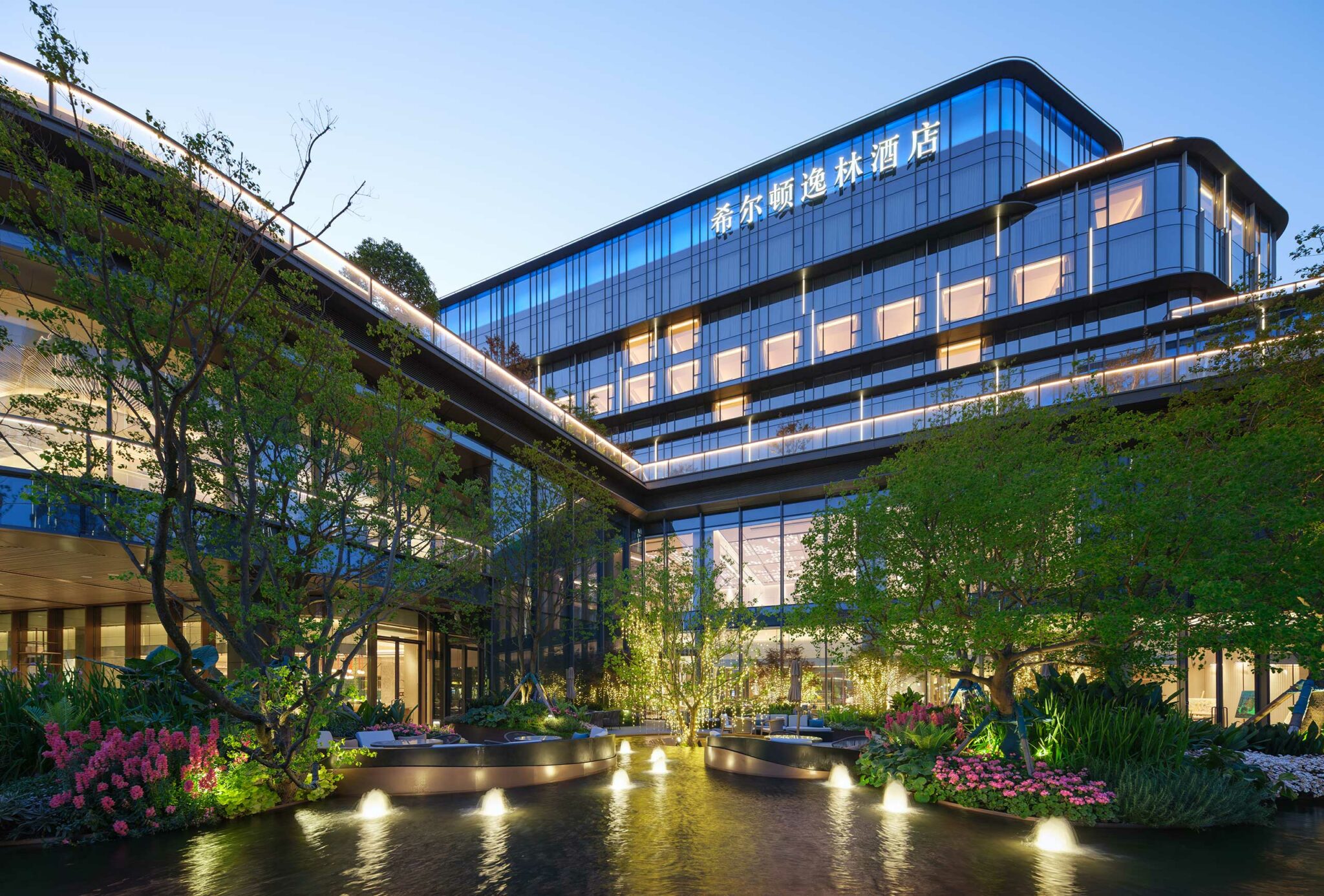 Modern hotel building with glass facade, surrounded by lush trees and flowering plants at dusk. A tranquil pond with lit fountains creates a serene ambiance.