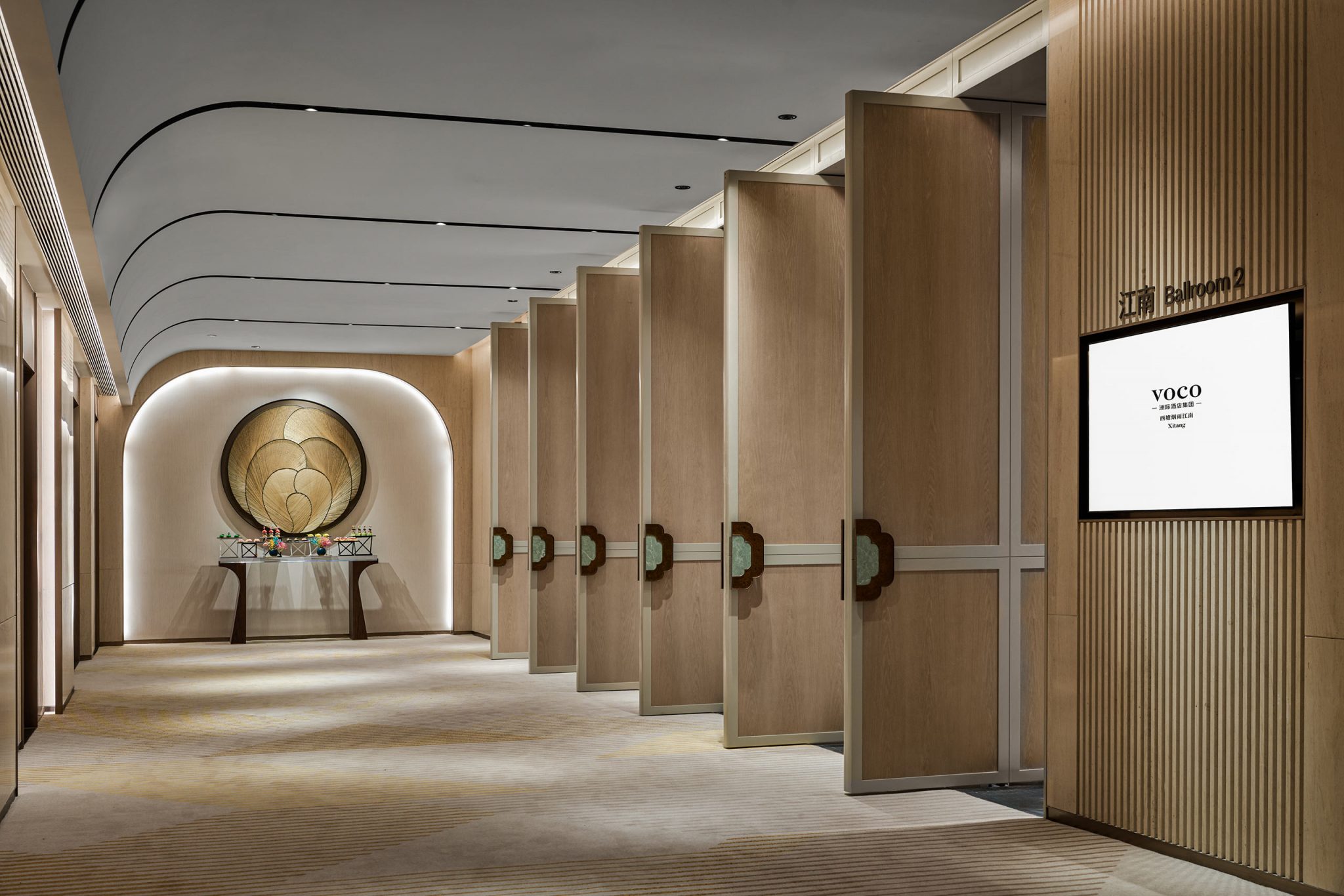 Modern pre-function hallway with beige walls and open wooden panel dividers leading to a bright room. An ornate wall decoration and table with glasses are seen.