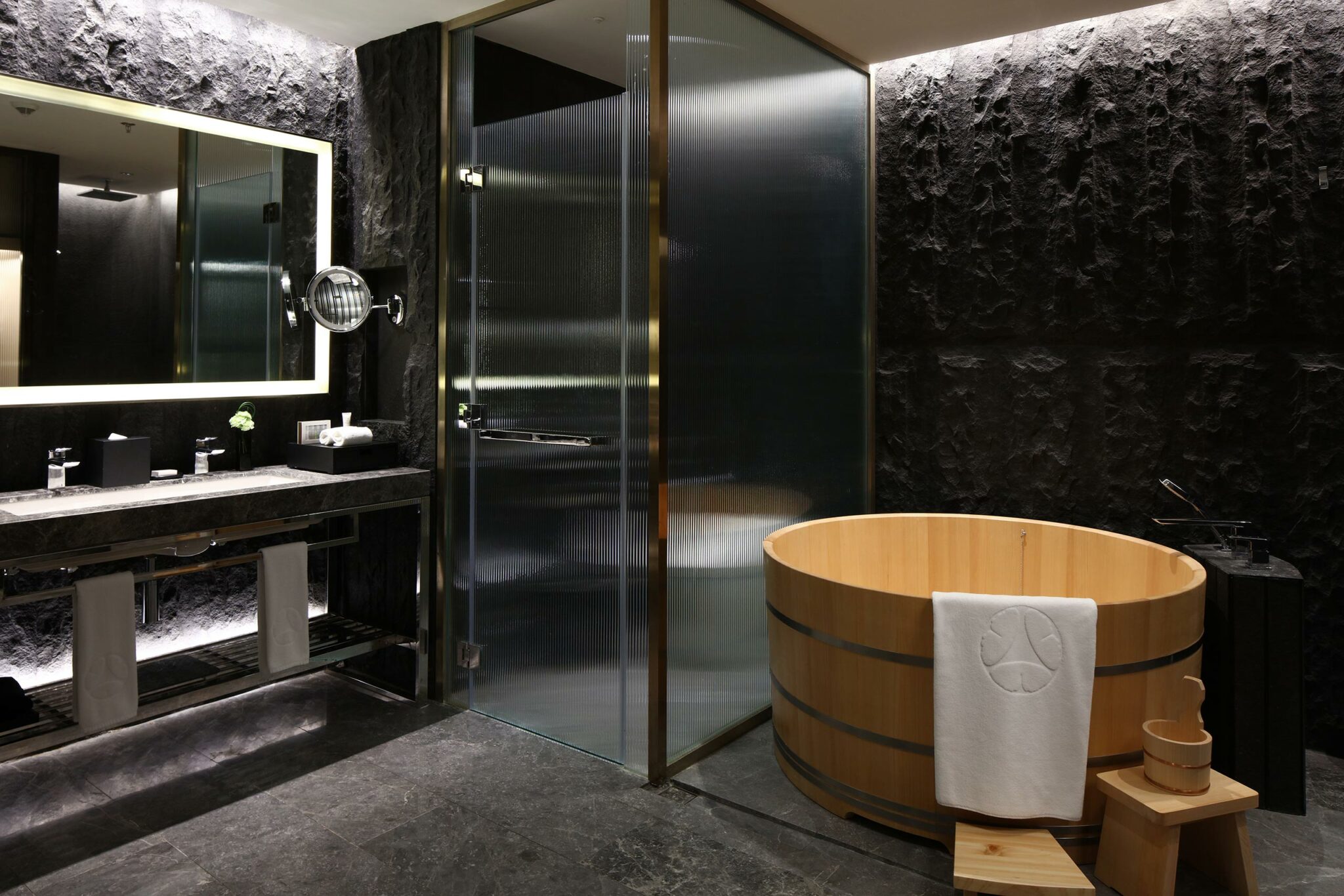 Luxurious guest room bathroom with dark stone walls, backlit mirror, glass shower, and wooden soaking tub. Minimalist and serene atmosphere.