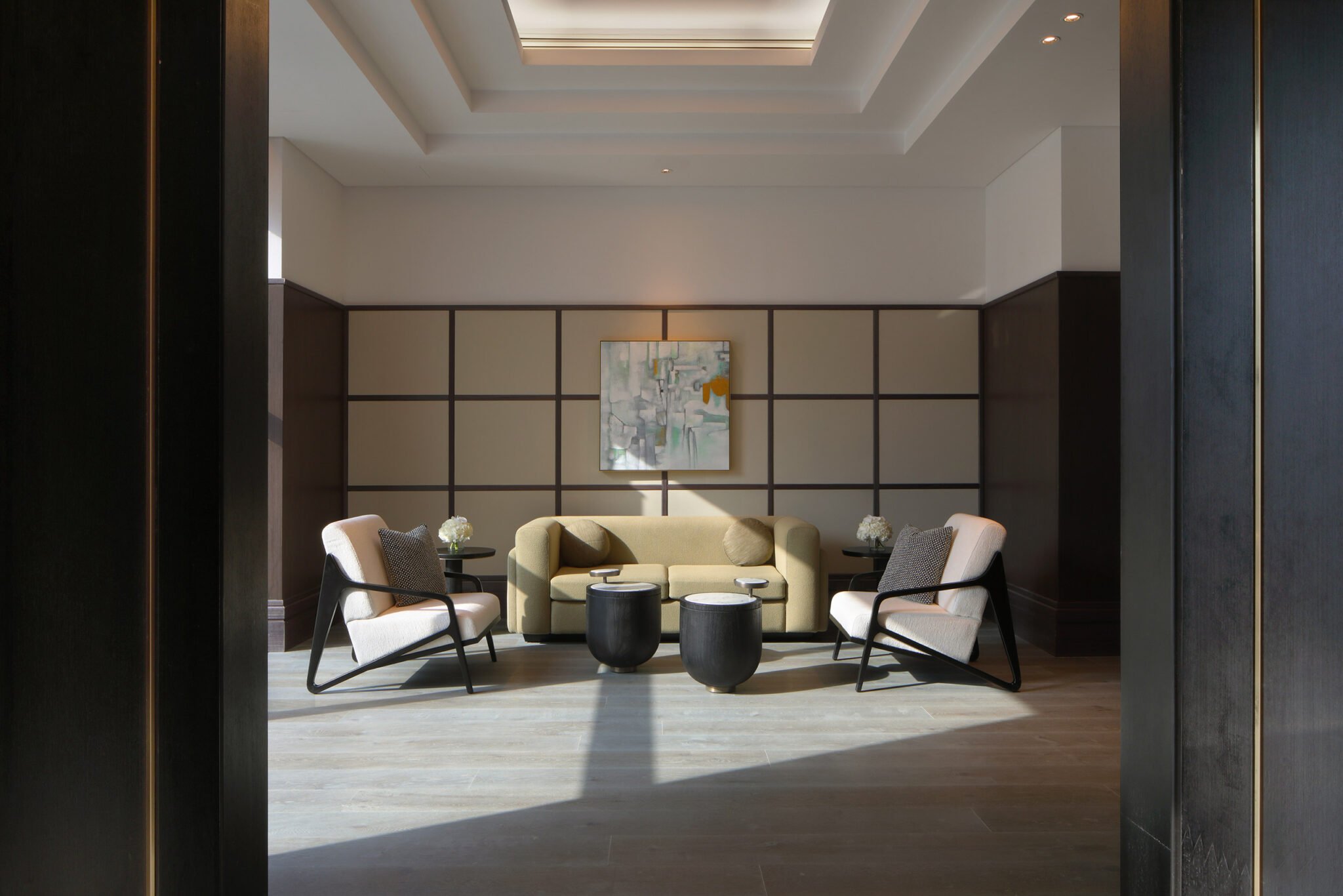 Modern hotel lounge with a beige sofa, two light armchairs, and black side tables. Abstract art hangs on a paneled wall, creating a calm, elegant ambiance.
