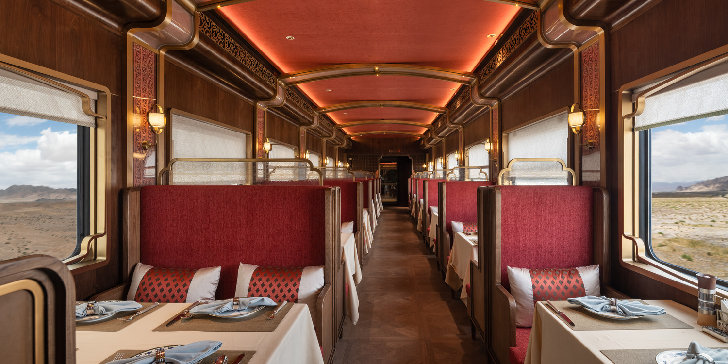 Luxurious train interior with elegant wooden paneling and large windows showing mountain views. Plush seating with decorative pillows creates a cozy atmosphere.