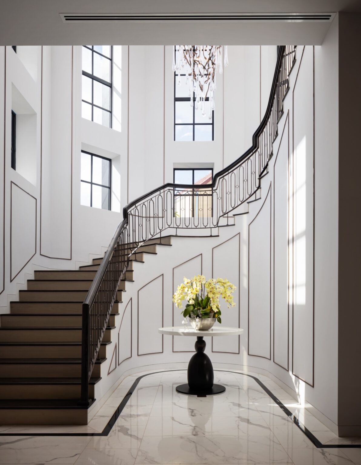 Elegant foyer with a grand staircase featuring dark wooden steps and sleek metal railing. Tall windows bring light in, illuminating an orchid on a central round table, creating a sophisticated atmosphere.