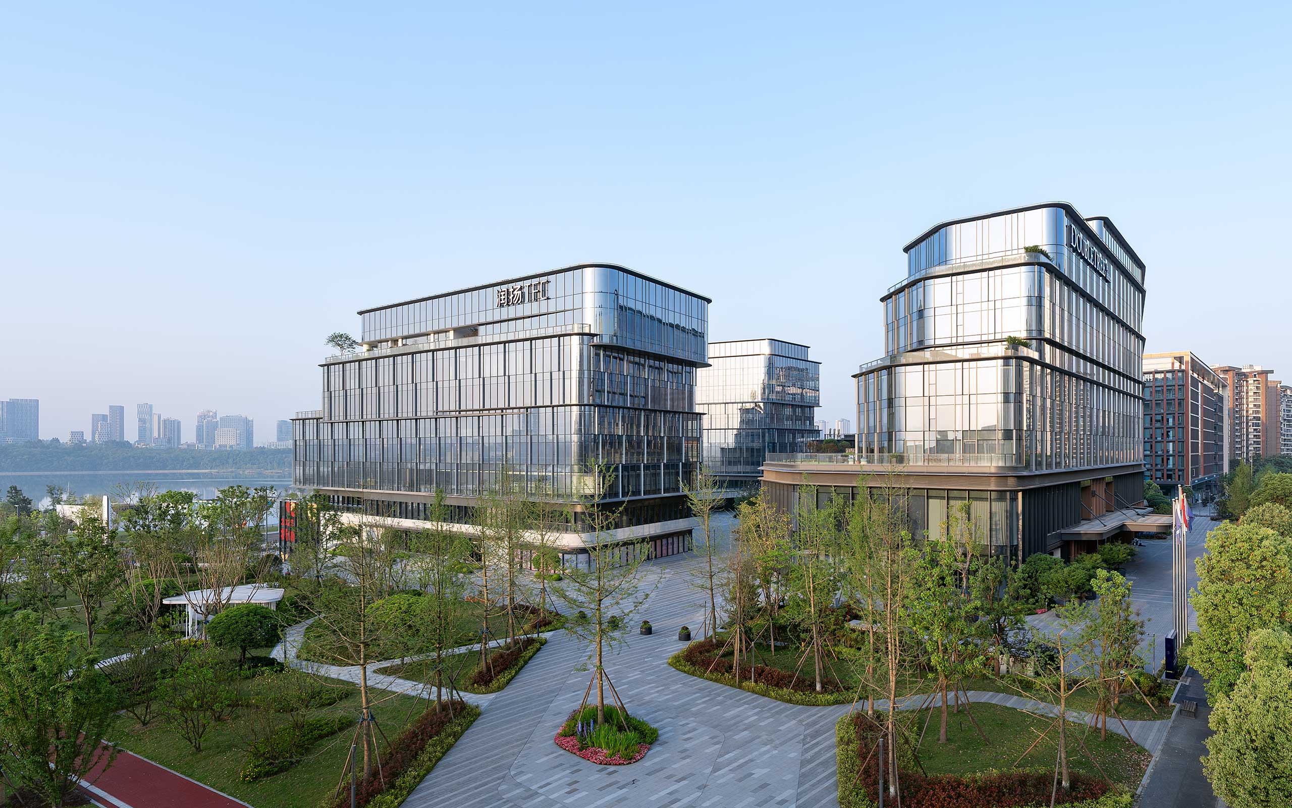 Modern commercial buildings with glass facades and rooftop gardens are surrounded by neatly landscaped greenery. The sky is clear and blue, creating a serene atmosphere.