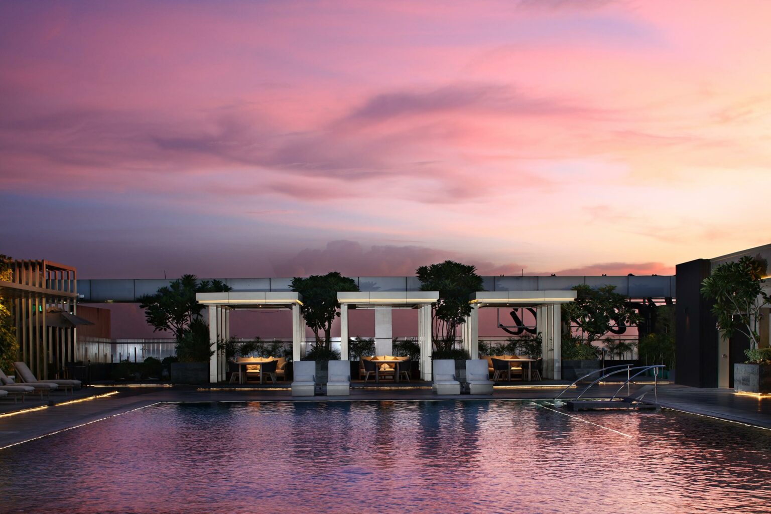 Rooftop pool at sunset with a pink and purple sky. Tables and chairs under pergolas surrounded by plants create a serene and inviting atmosphere.