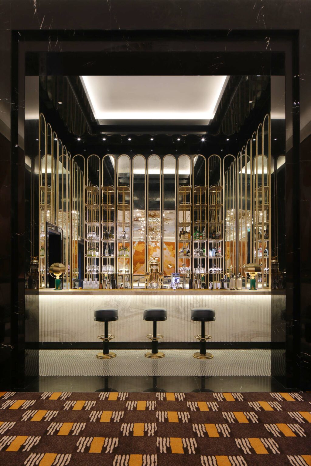 Luxurious hotel bar with gold accents, a reflective ceiling, and elegant lighting. Three black stools line the bar, conveying a sleek, sophisticated ambiance.