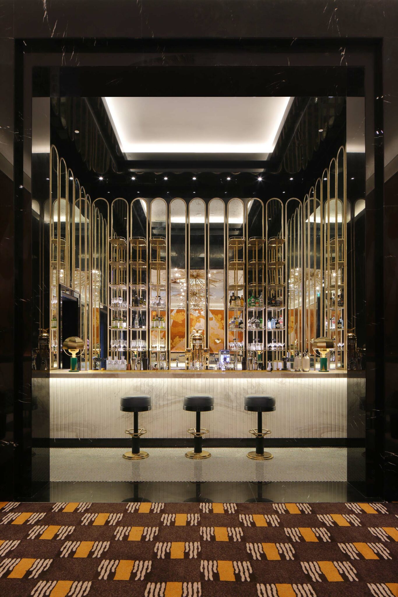 Luxurious hotel bar with gold accents, a reflective ceiling, and elegant lighting. Three black stools line the bar, conveying a sleek, sophisticated ambiance.