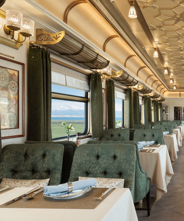 Luxurious train interior with elegant wooden paneling and large windows showing mountain views. Plush seating with decorative pillows creates a cozy atmosphere.