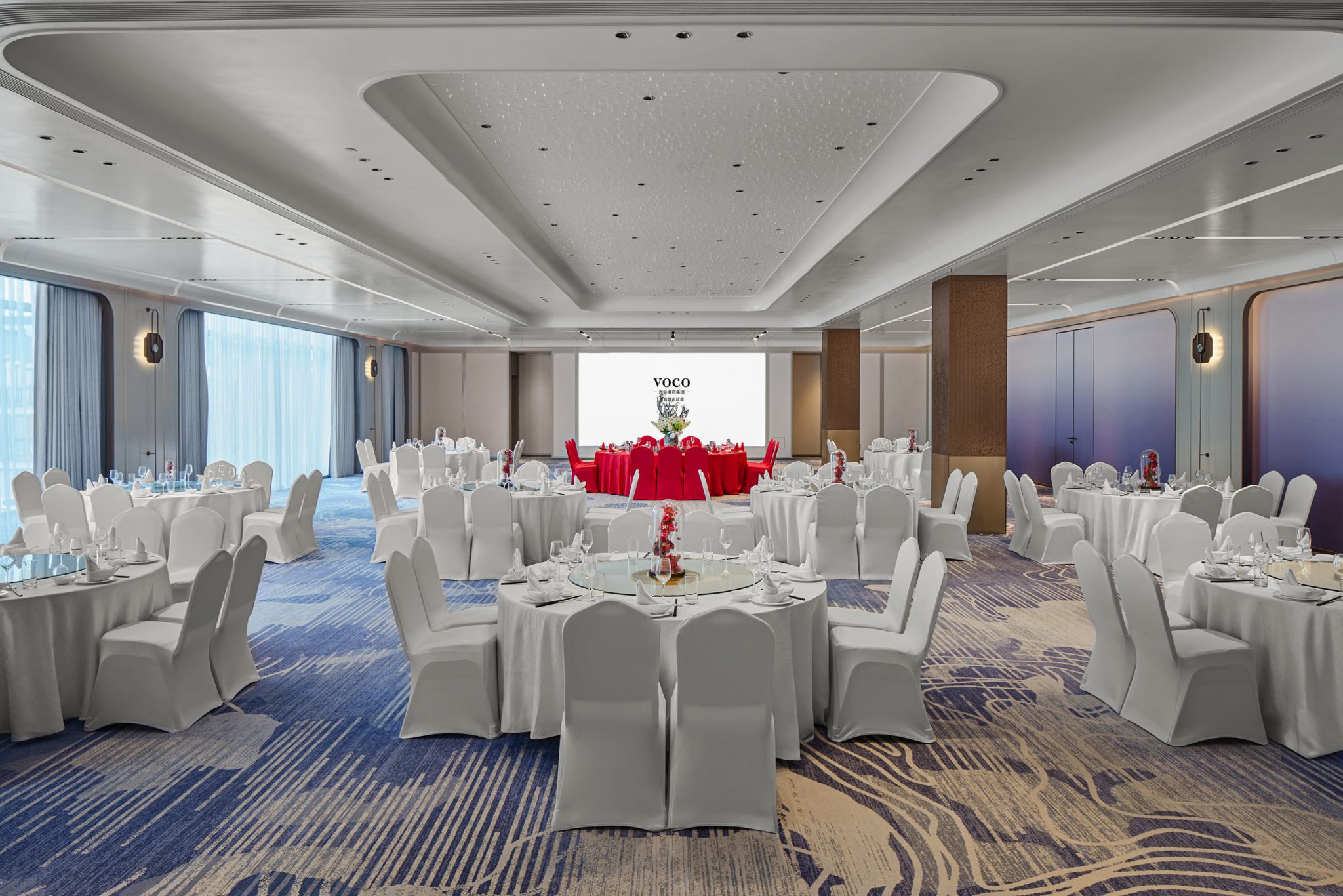 Elegant banquet hall with round tables draped in white linens, contrasting with bright red tablecloths at the front. Soft lighting and cozy ambiance.