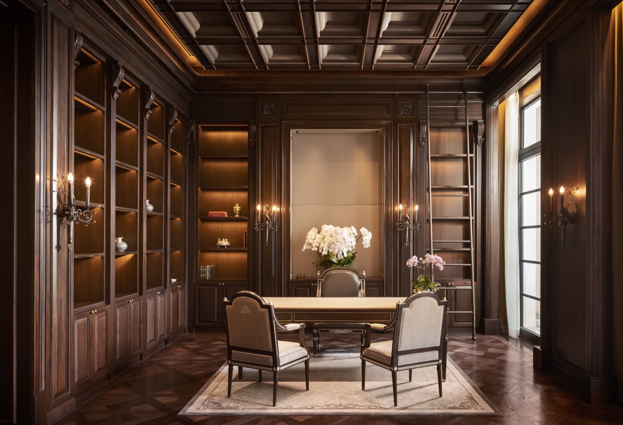 Elegant study with dark wooden shelves, ladder, and orchids on a central table. Soft lighting from wall sconces provides a warm, sophisticated ambiance.