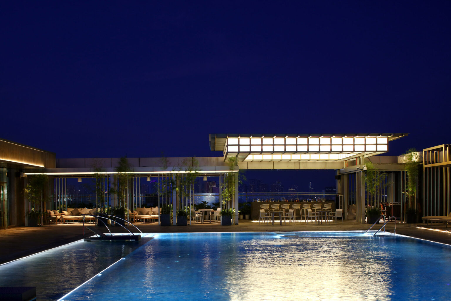 A serene rooftop pool at night, bordered by bamboo plants. Soft lighting illuminates the water and nearby seating area, evoking a tranquil ambiance.