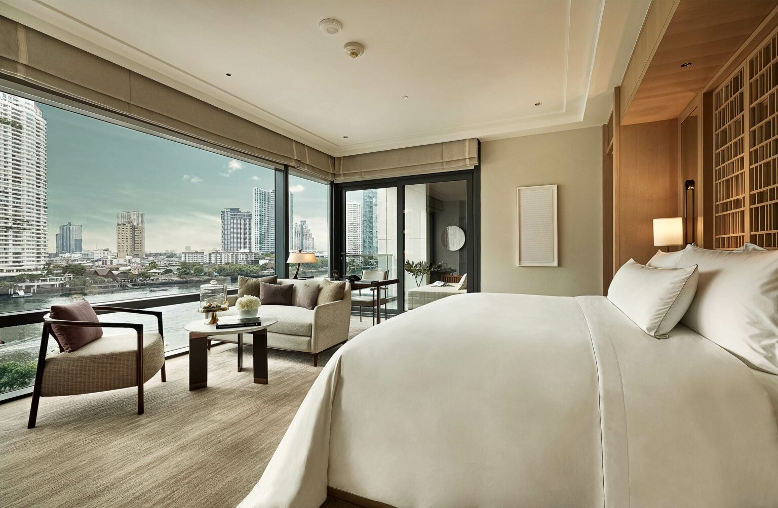 Luxurious hotel room with a white bed and large windows showcasing Bangkok's skyline. Soft lighting, elegant furniture, and a serene ambiance.