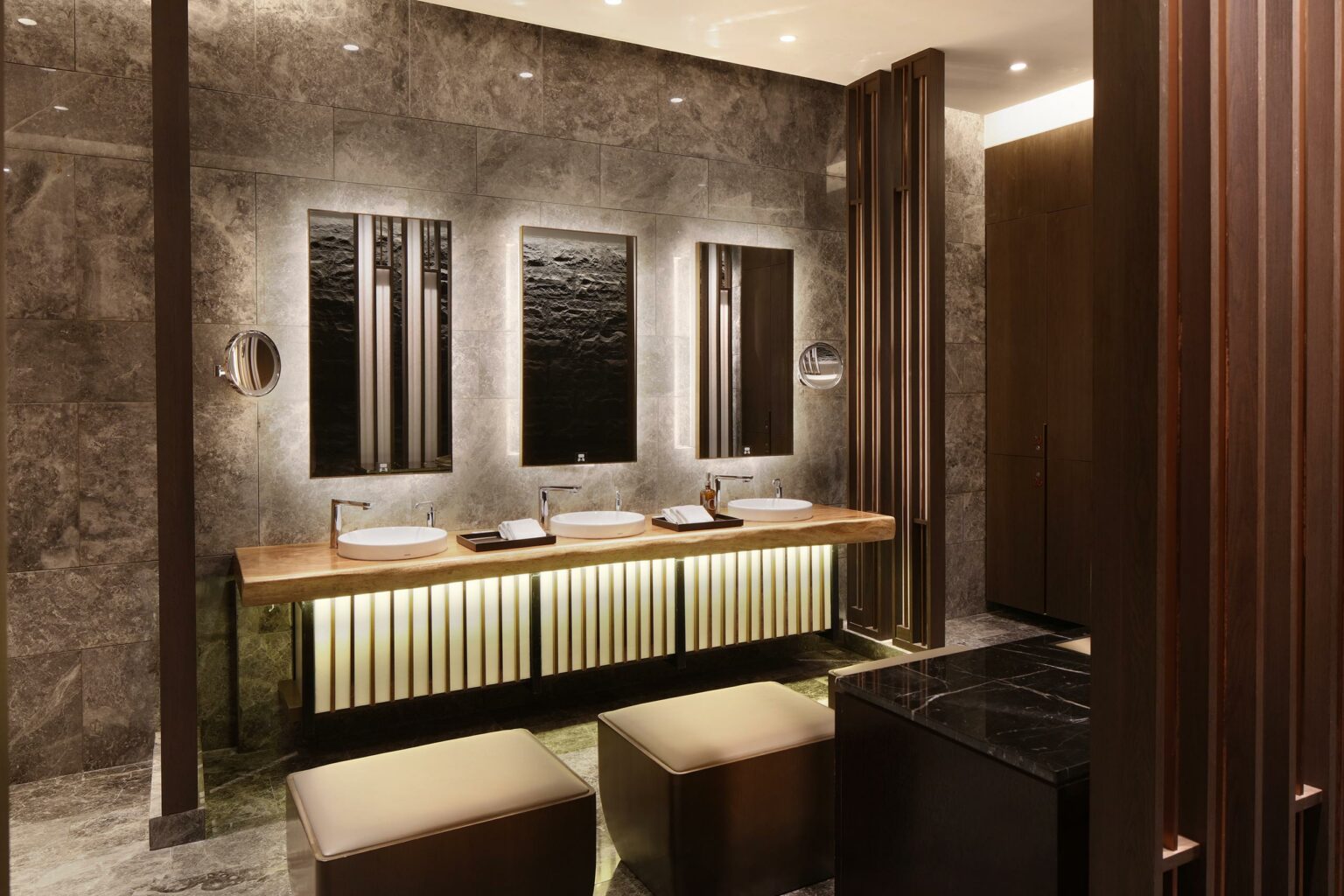 Luxurious hotel changing room with three backlit mirrors, round sinks on a wooden counter, and warm ambient lighting. Stone walls and elegant minimalist decor.