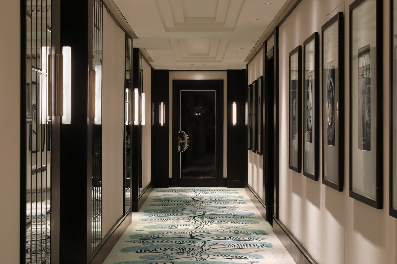 Elegant hotel corridor featuring soft lighting, a patterned carpet, and framed art lining the walls, leading to a dark, closed door. Quiet and refined ambiance.