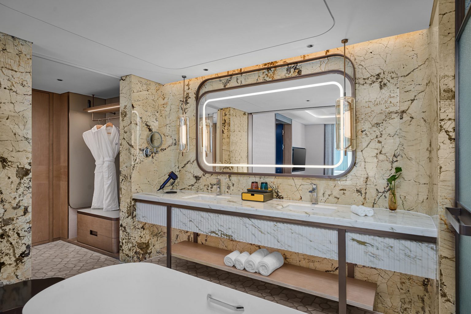 Luxurious bathroom with marble walls, large illuminated mirror, dual sinks on a sleek counter, rolled towels beneath, and a robe hanging by a closet.