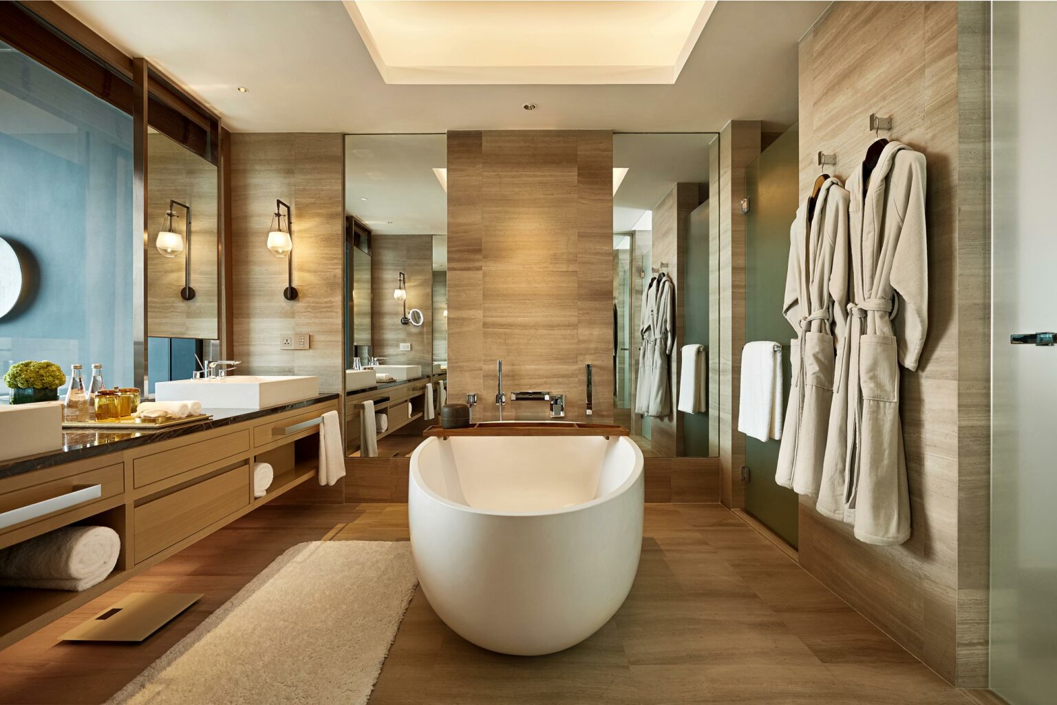 Luxurious hotel bathroom with a freestanding oval tub, wood accents, and wall mirrors. Soft lighting, plush robes, and towels create a serene, spa-like atmosphere.