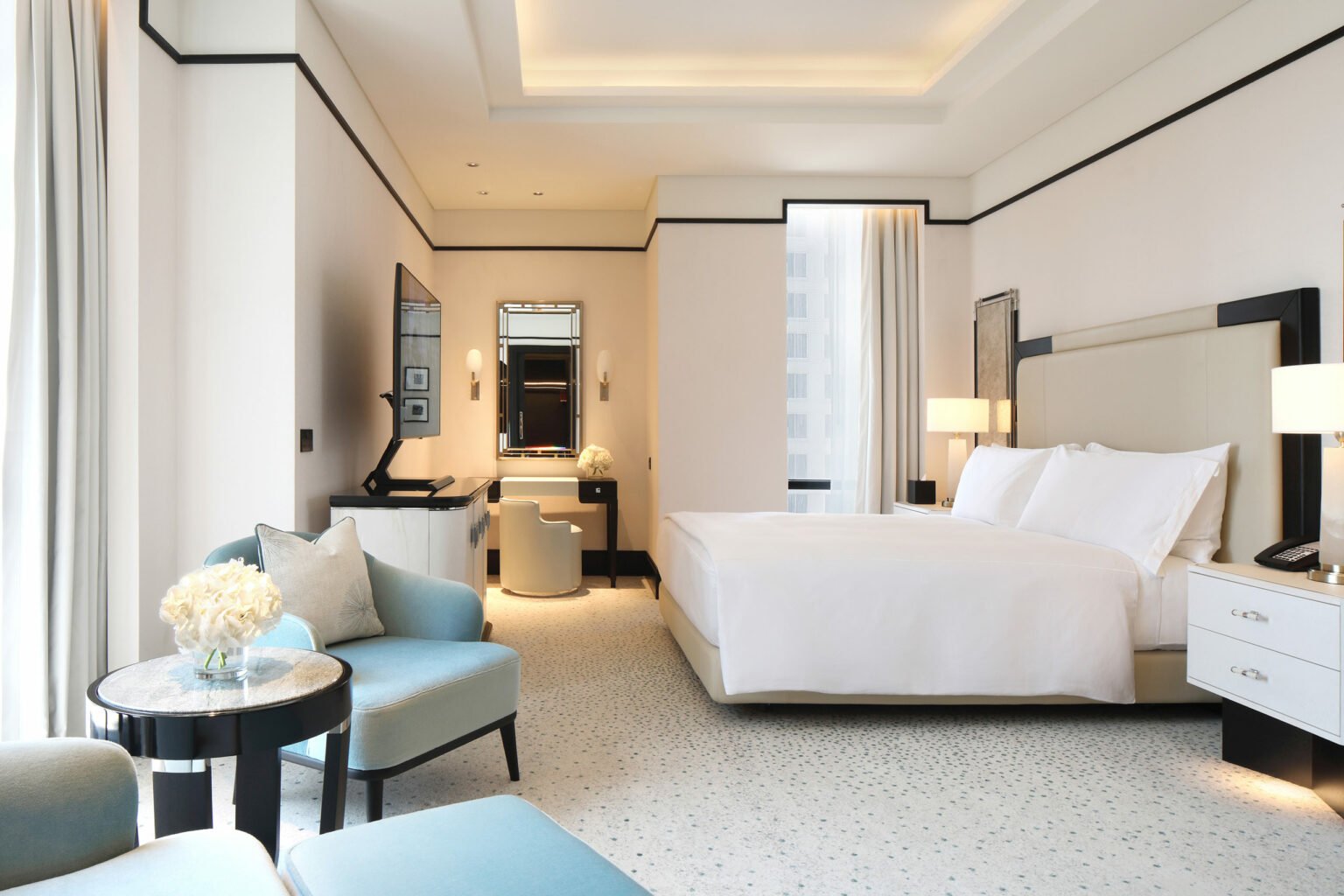 A luxurious hotel room features a large bed with white linens, elegant furniture, soft lighting, and a serene ambiance. Light blue chairs face a small table with flowers.