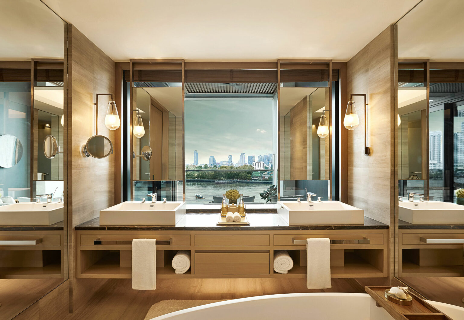 Luxurious hotel bathroom with two sinks, wooden cabinets, and large mirrors. A window offers a skyline view of Bangkok, creating a serene, elegant ambiance.