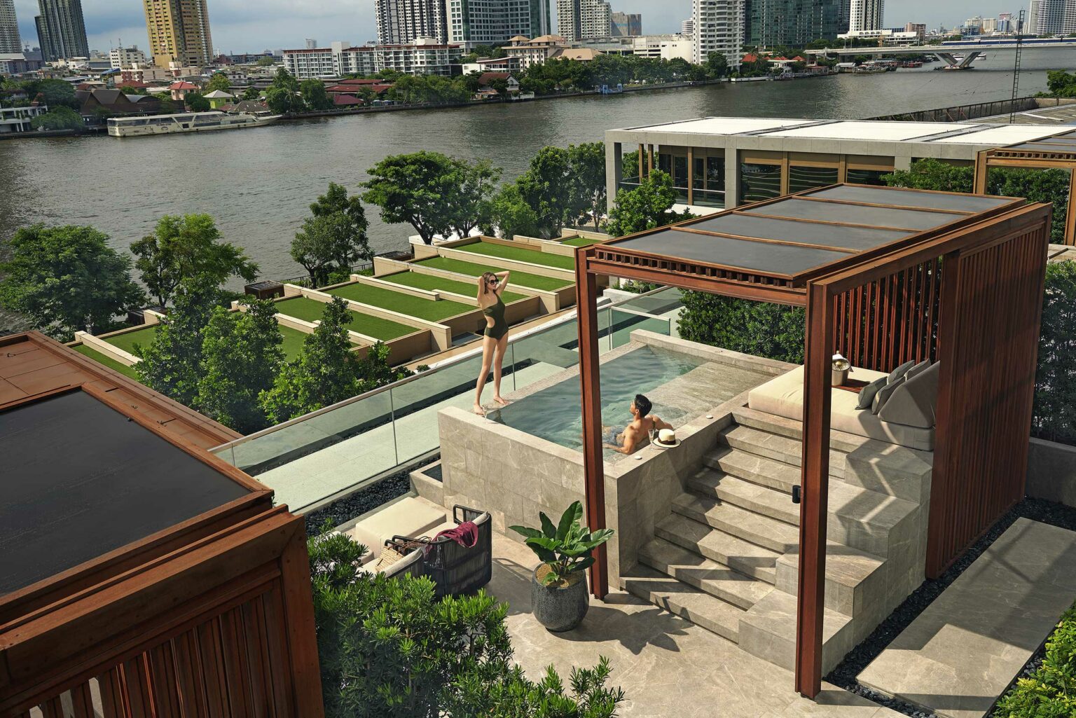 Hotel rooftop patio with a small pool overlooks a river. One person stands outside, another relaxes in the pool under a wooden pergola. Lush greenery surrounds.