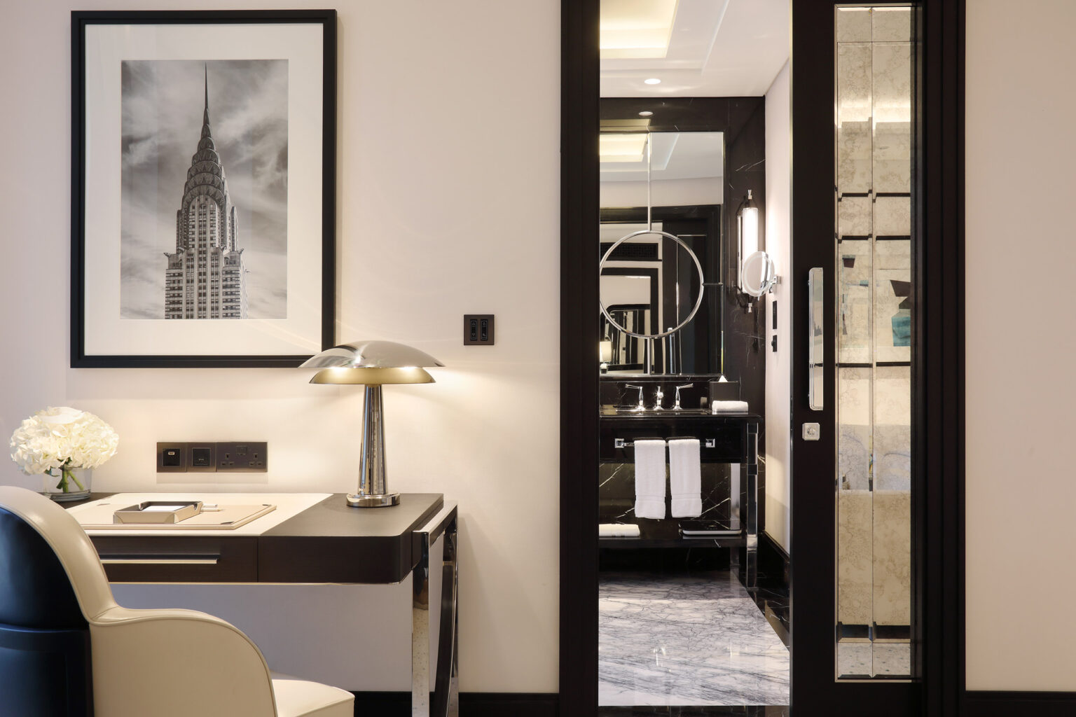 Modern suite; black and white cityscape photo, sleek desk with lamp, elegant adjoining bathroom with towels, reflecting luxury and sophistication.