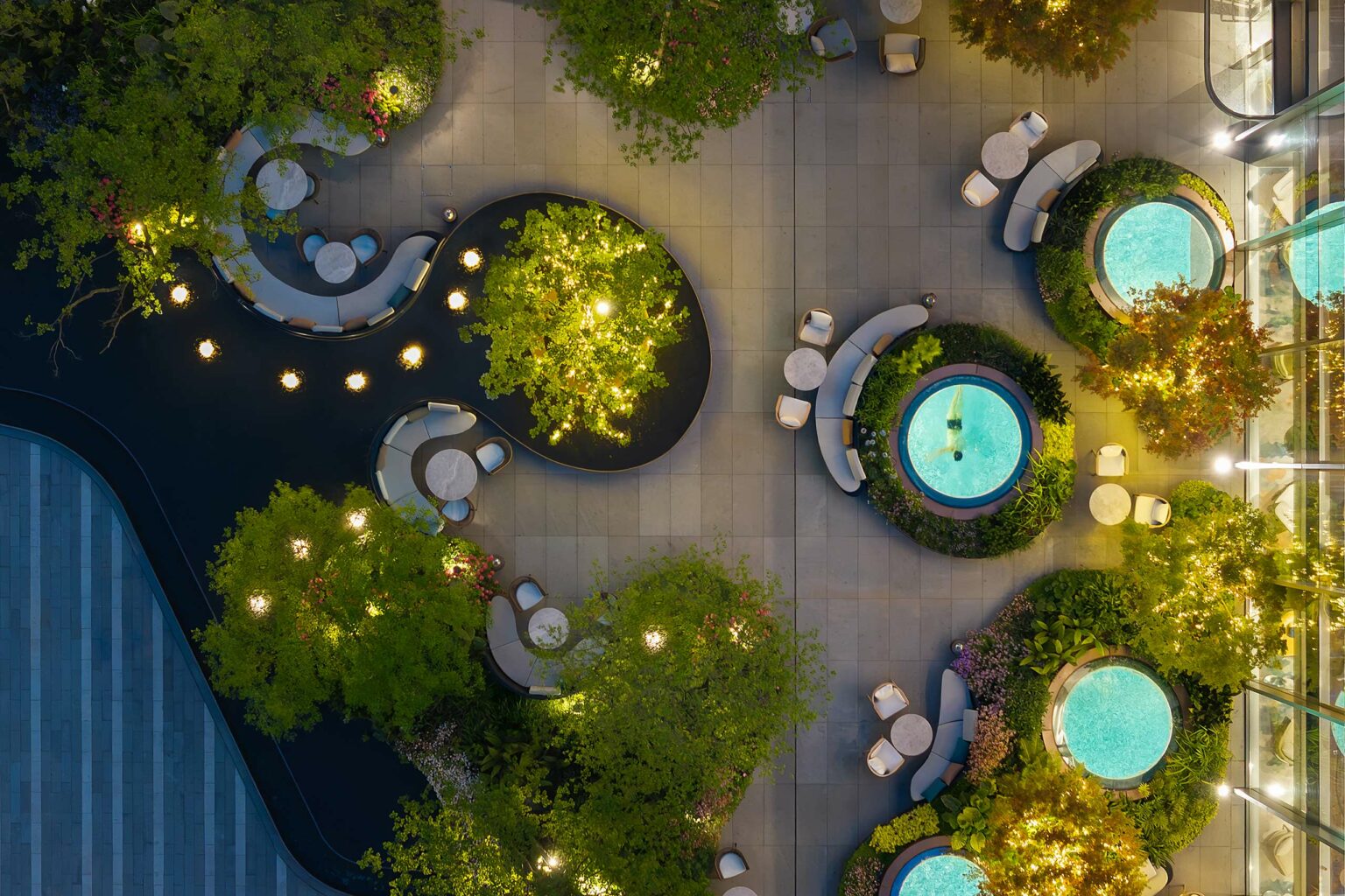 Aerial view of a modern outdoor terrace at night with circular seating areas and tables. Pools with turquoise lighting are surrounded by lush greenery.