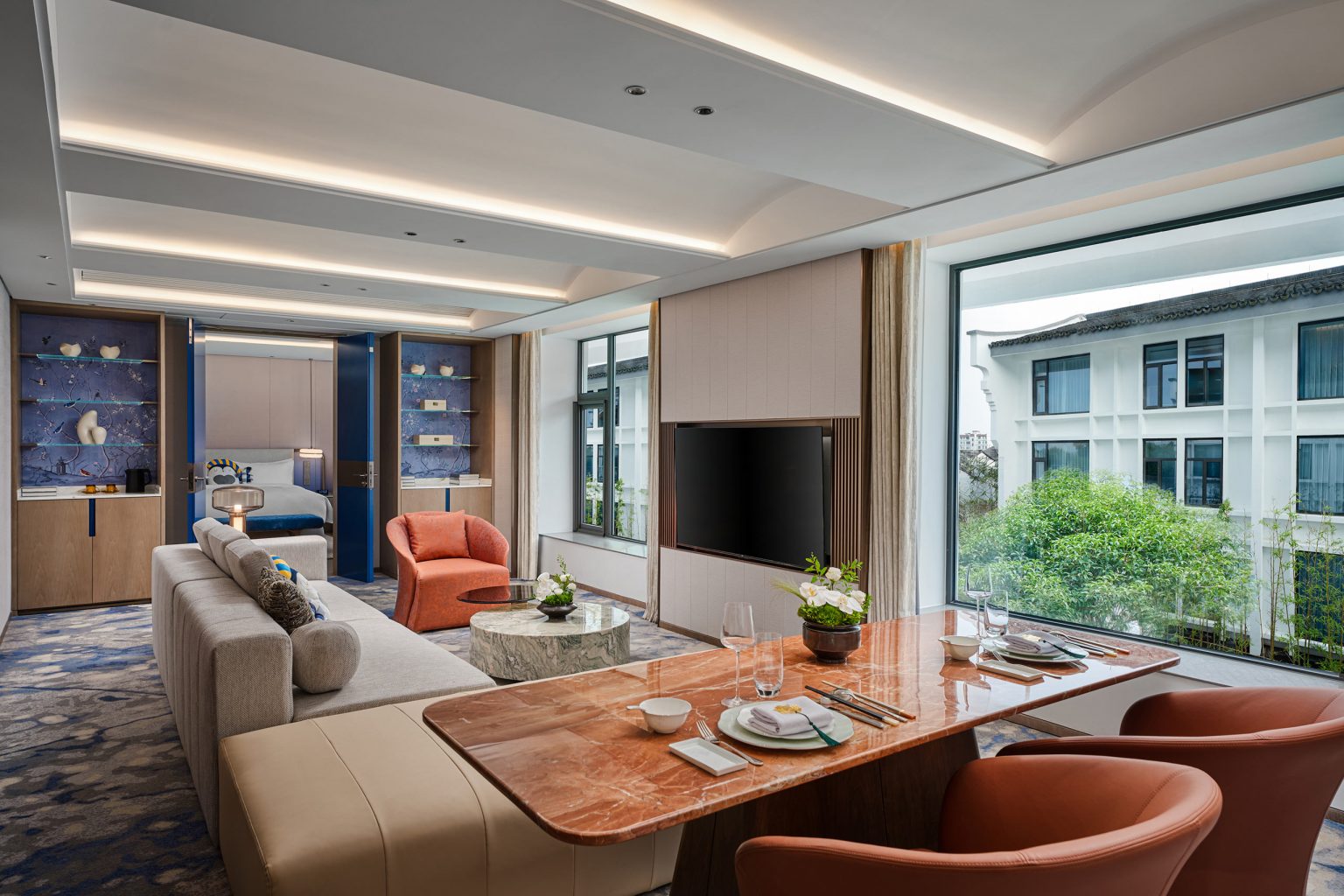 Modern living room with a large window view, featuring a beige sofa, orange chairs, marble dining table, and built-in shelving, creating a cozy ambiance.