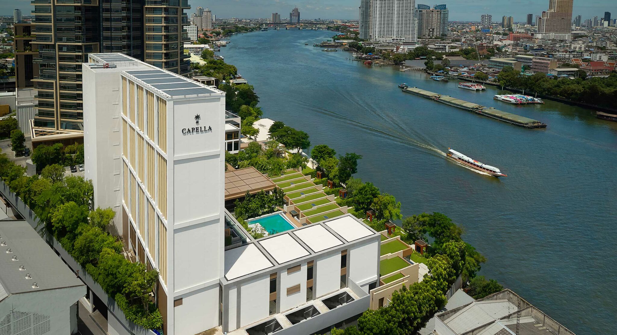 Aerial view of Capella Bangkok, featuring a modern riverside hotel with lush greenery, a pool, and manicured lawns. A boat glides on the river, set against a city backdrop.