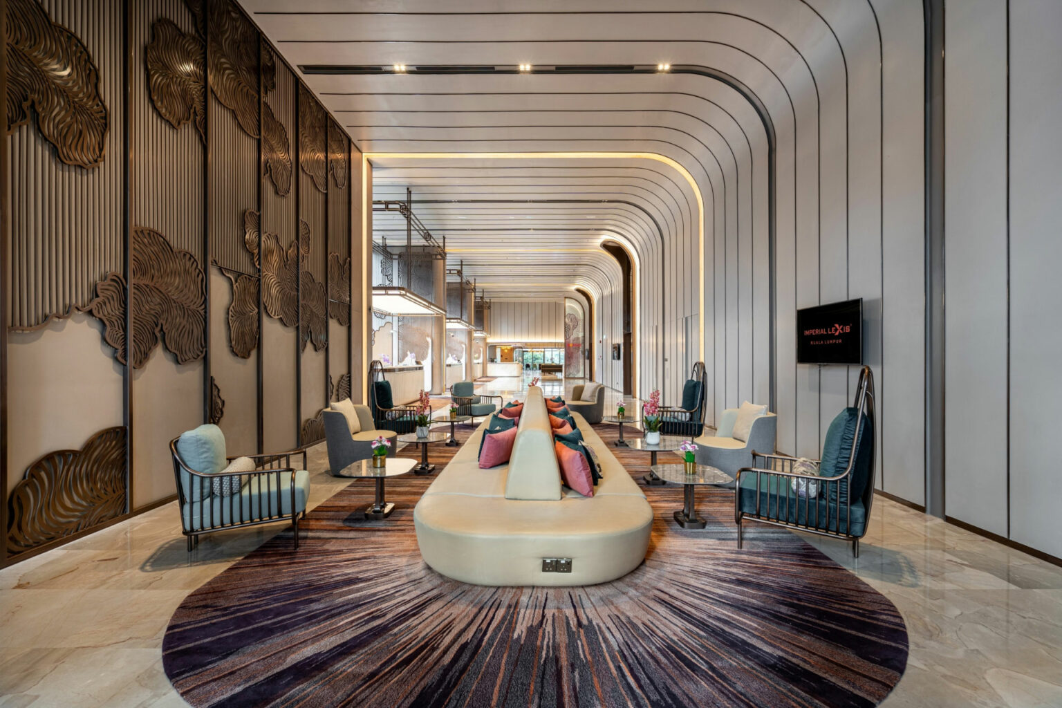 Modern hotel lobby with high, arched ceiling. Cream seating, colorful pillows, and elegant chairs line a central aisle. Decorative walls and warm lighting create a luxurious, inviting atmosphere.