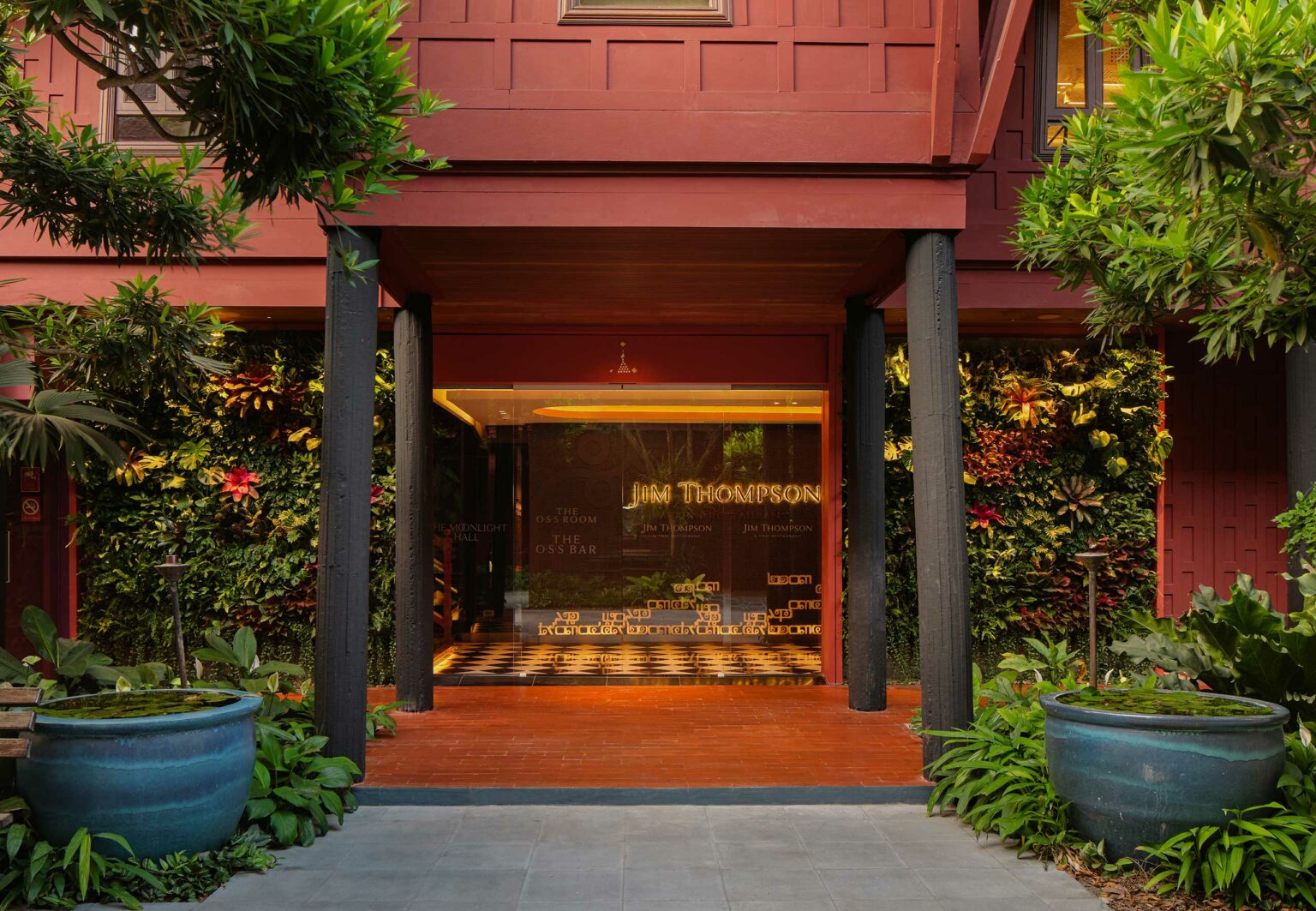 Entrance of Jim Thompson Restaurant Bangkok with red walls and black pillars, surrounded by lush greenery and painted pots. Glass doors display 