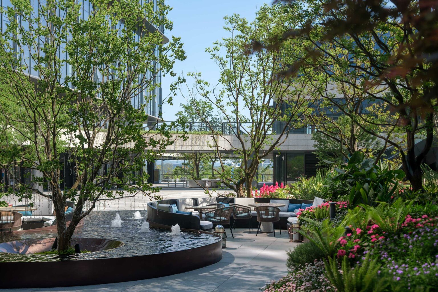 Modern outdoor terrace, featuring a curved water fountain, lush greenery, and seating area with wicker chairs and sofas, creating a serene ambiance.
