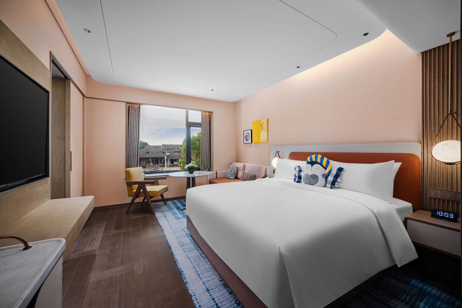 Modern hotel room with a large bed, blue bench, and plush pillows. Elegant lighting and a luxurious bathroom with a tub add sophistication and tranquility.