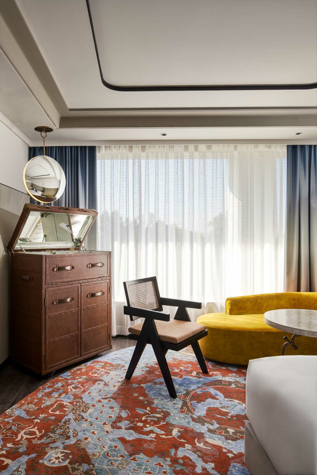 Modern hotel room with a cozy yellow sofa, round table, and vintage dresser. A large bed is in the foreground. Soft light filters through sheer curtains.