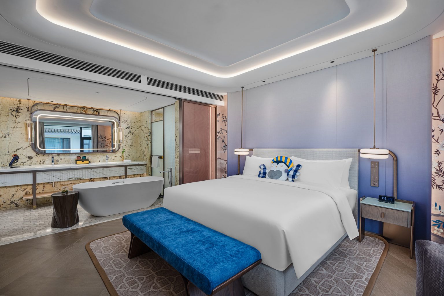 Modern hotel room with a large bed, blue bench, and plush pillows. Elegant lighting and a luxurious bathroom with a tub add sophistication and tranquility.