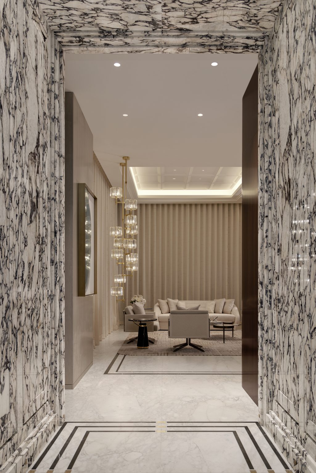 Marble hallway opens to a modern living room with a beige sofa, glass chandelier, side table, and striped wallpaper. Ceiling lights illuminate the space.