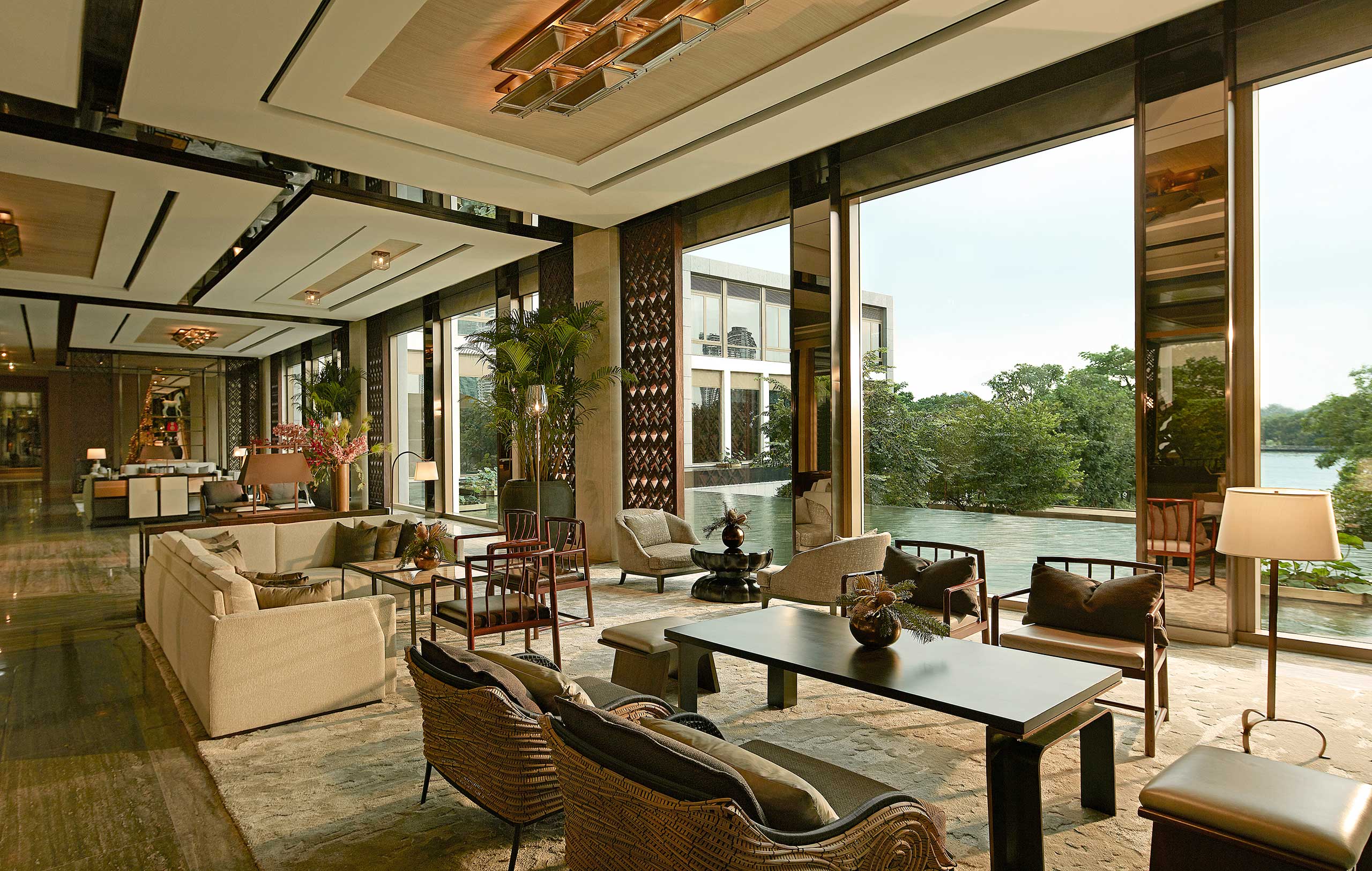 Luxurious hotel tea lounge with modern decor, beige sofas, and large windows overlooking a lush garden and water. Warm lighting creates an inviting atmosphere.