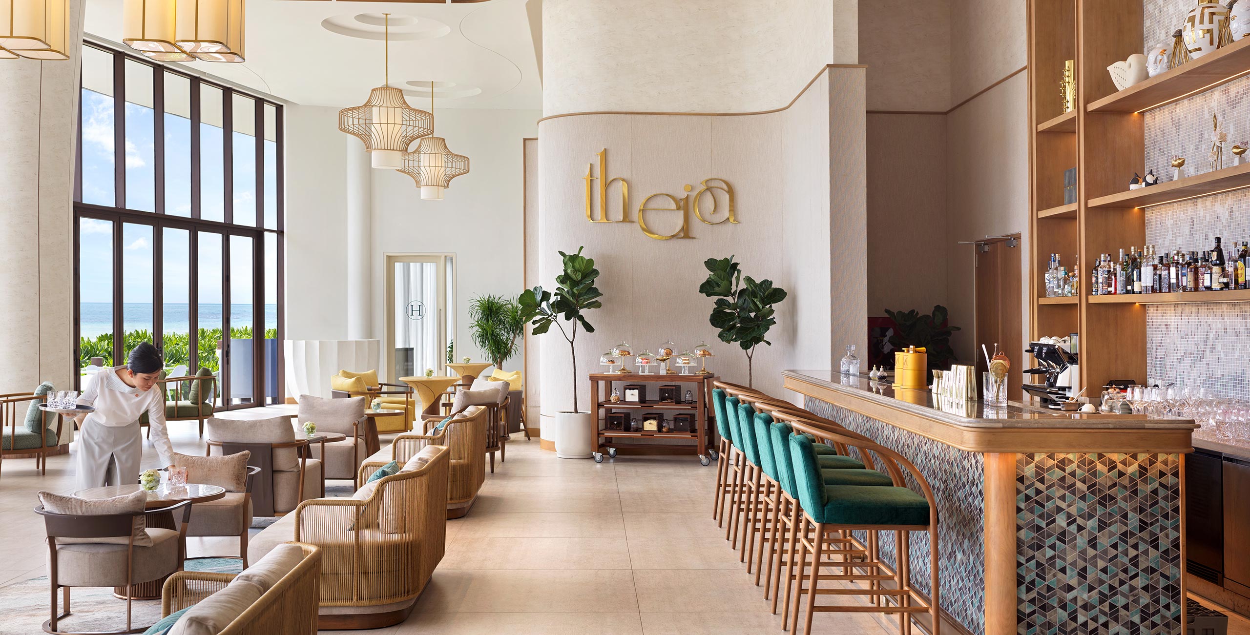 Luxurious, airy hotel cafe with ocean view. Features wicker chairs, green bar stools, potted plants, modern decor, and a server arranging items.