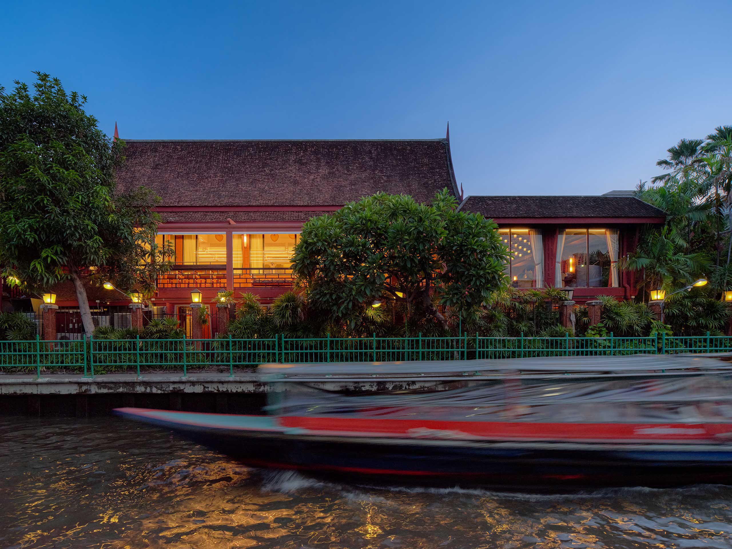Jim Thompson Restaurant Bangkok's traditional Thai building with warm lights and lush greenery, viewed from across a canal. A blurred boat speeds by under a twilight sky.