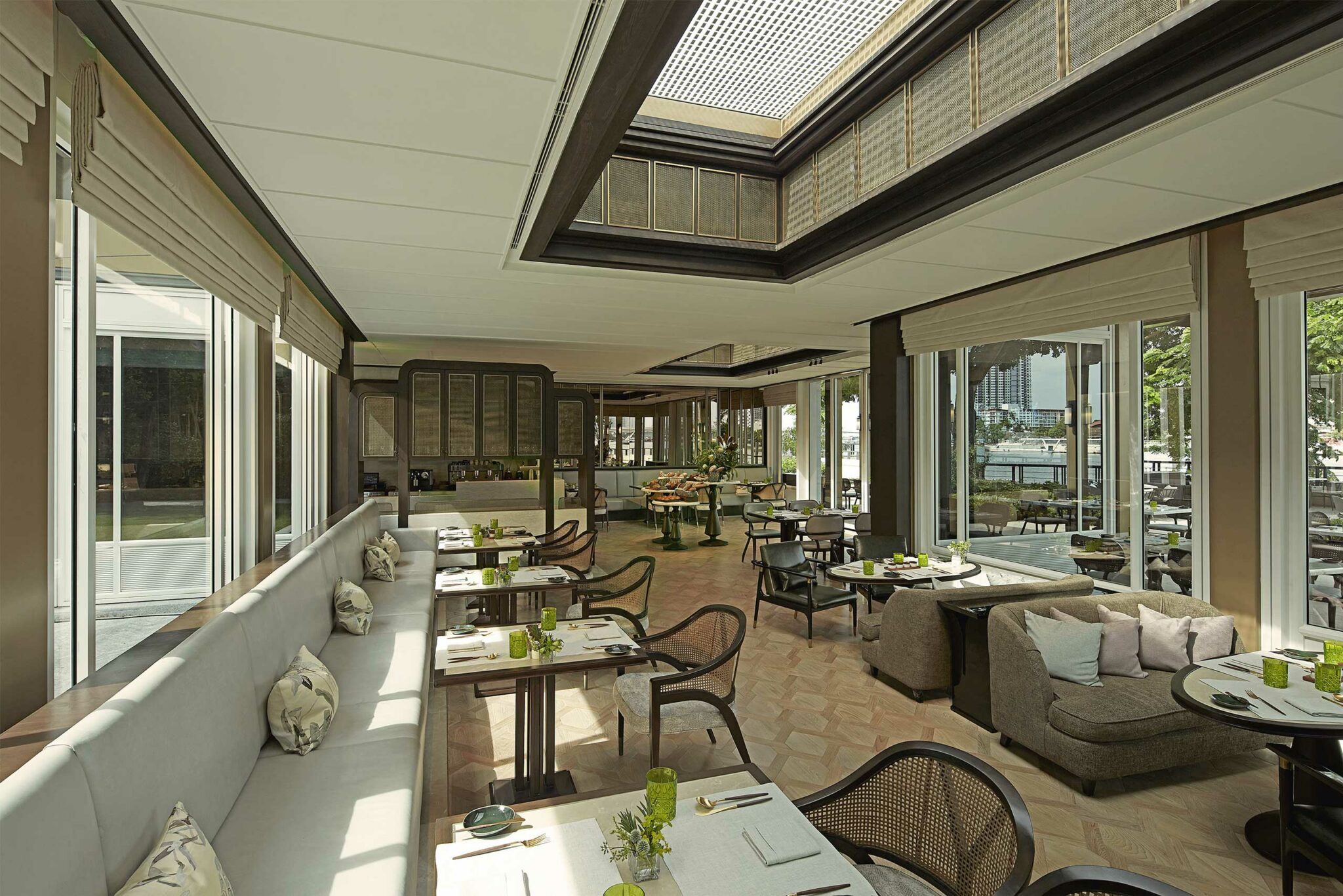 Bright, modern restaurant with large windows, elegant tables set for dining, and plush seating. Green accents and natural light create a cozy, inviting atmosphere.