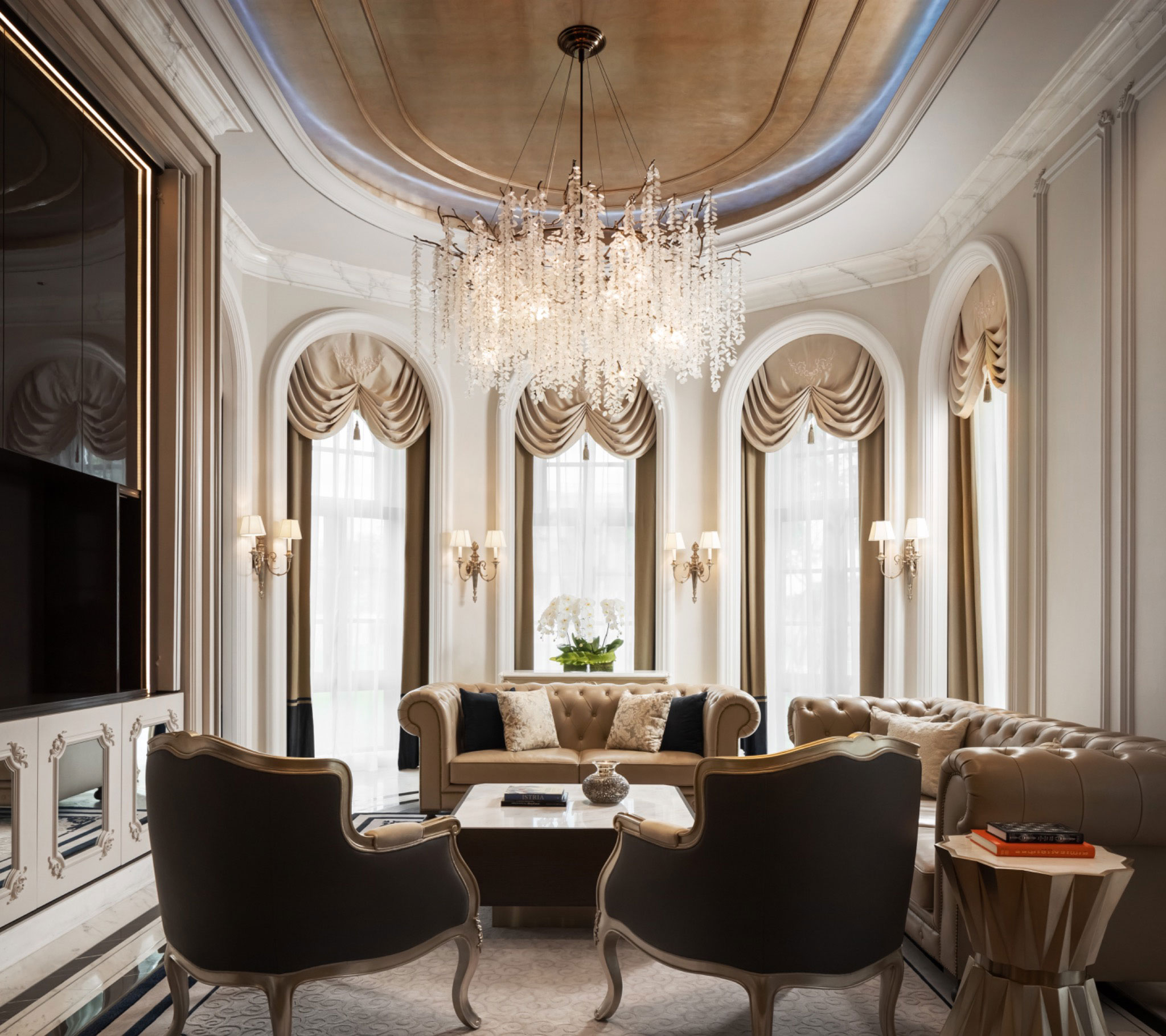 Elegant living room featuring a grand crystal chandelier, tufted sofas, arched windows with drapes, and a polished coffee table with a serene ambiance.