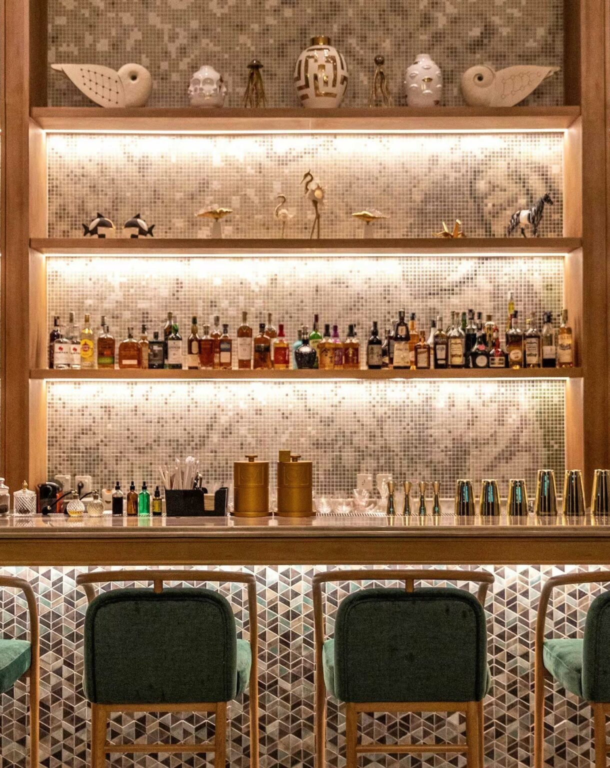 A stylish bar with teal chairs and a patterned counter. Shelves display liquor bottles and artistic decor, set against an elegant tile backdrop. Soft lighting creates a warm ambiance.