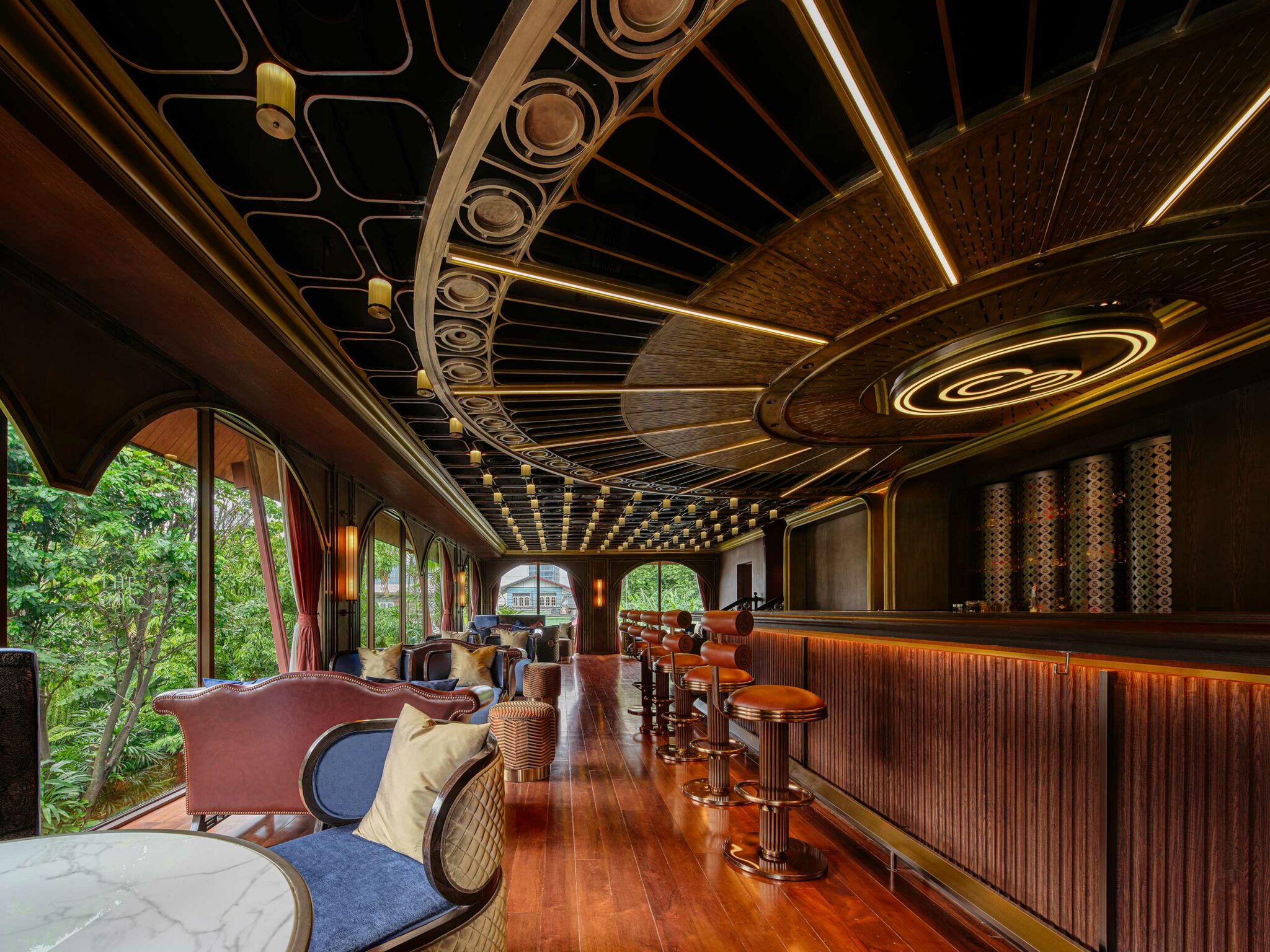 Luxurious restaurant bar area with wooden floors and curved, art deco ceiling. Tall bar stools line the counter, with plush seating by large windows showing greenery.