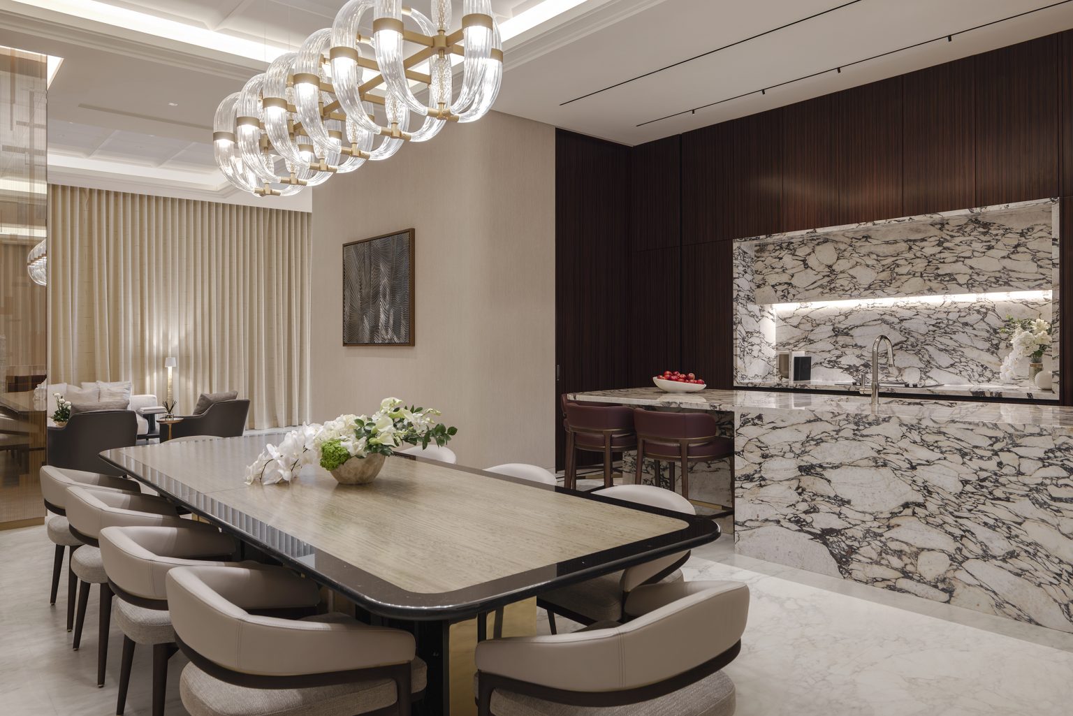 Elegant dining room of Waldorf Astoria Residences Dubai with a long, wooden table surrounded by cushioned chairs. A modern chandelier hangs above. Marble kitchen counter and cozy seating area.